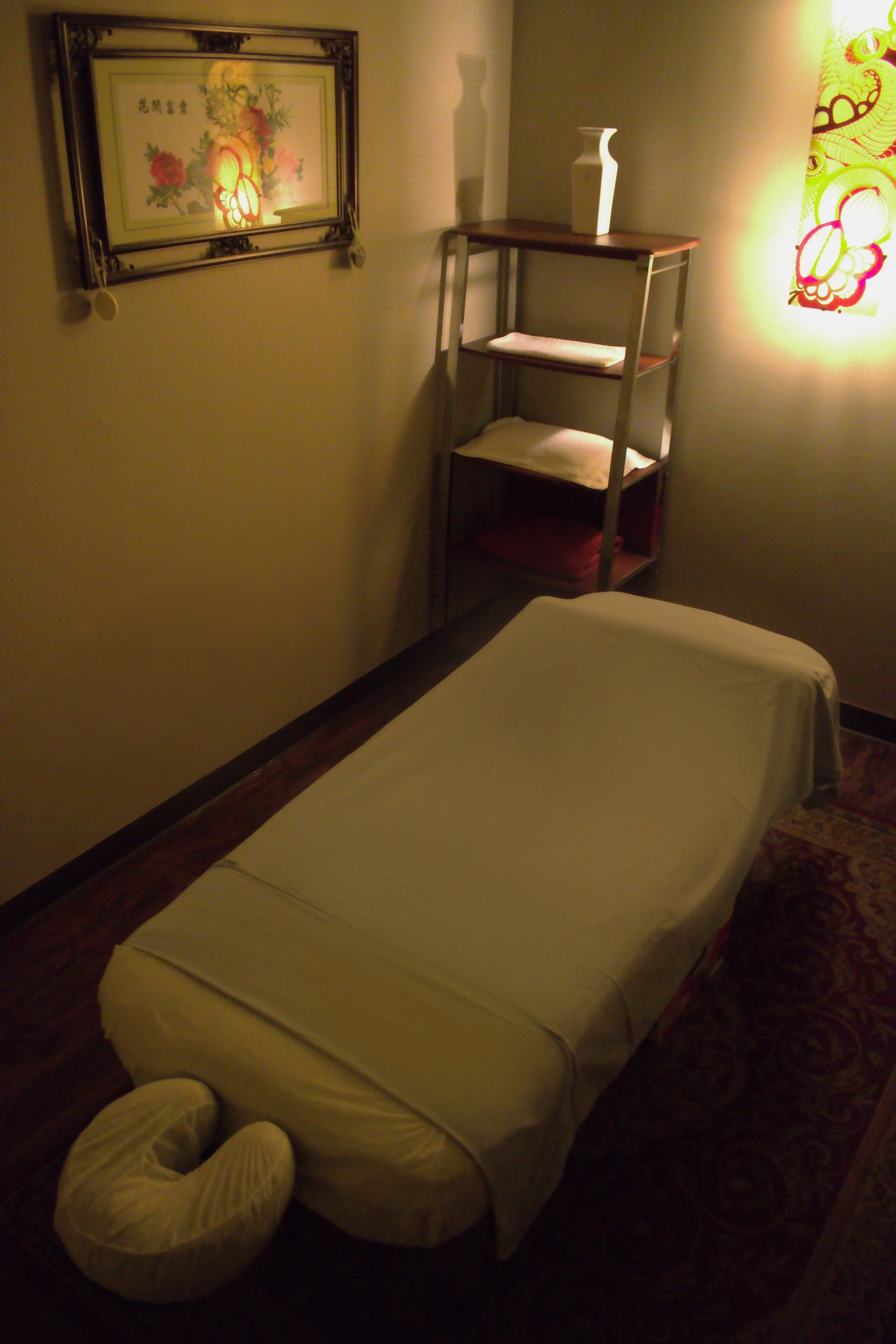 A massage table covered with a white sheet in a dimly lit room, with a neck pillow at one end. A wooden shelf with folded towels and a white vase is against the wall. Decorative art and a framed picture hang on the walls.