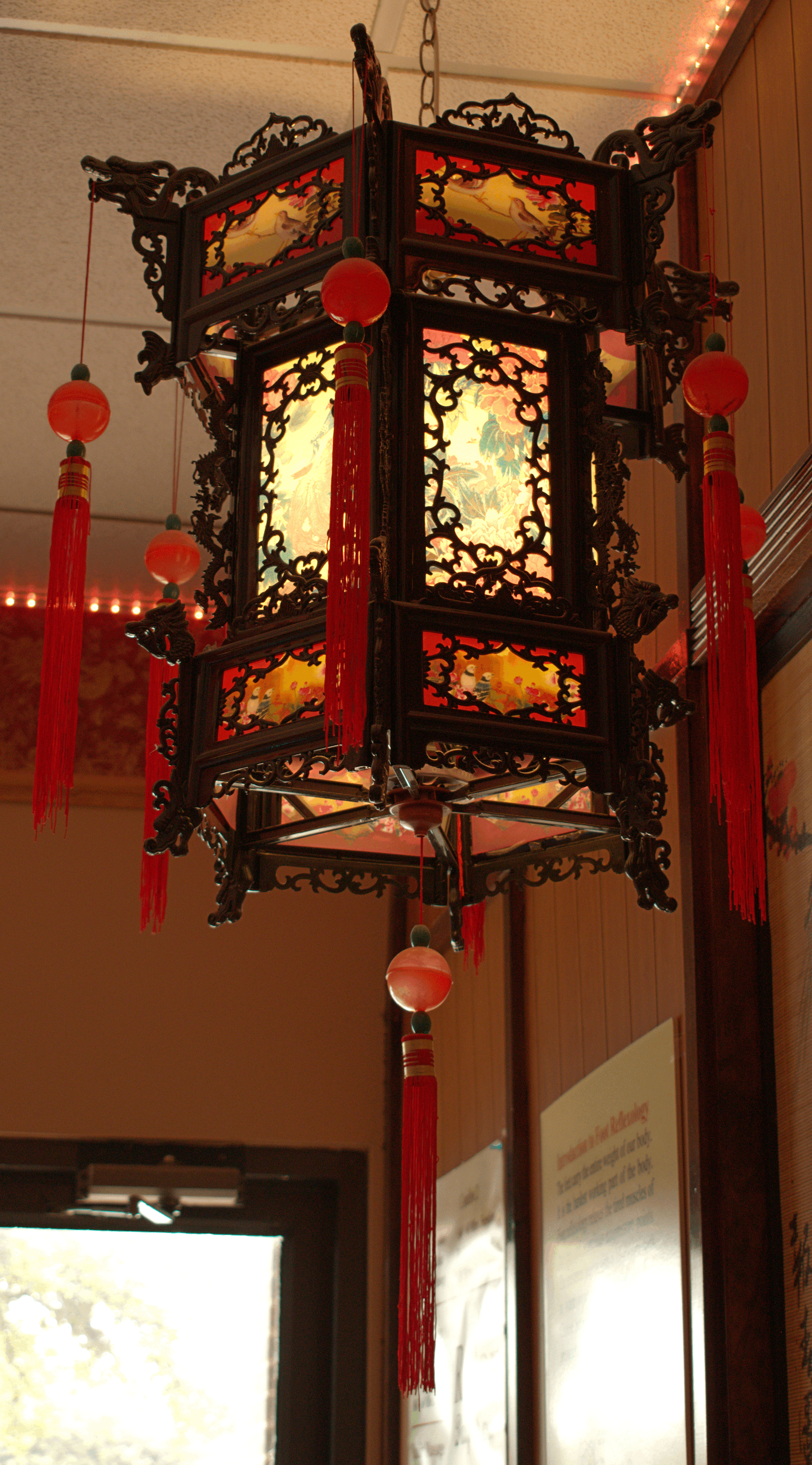 A traditional Chinese hanging lantern with intricate wooden carvings and red tassels, illuminated from within, hanging from the ceiling in a room.