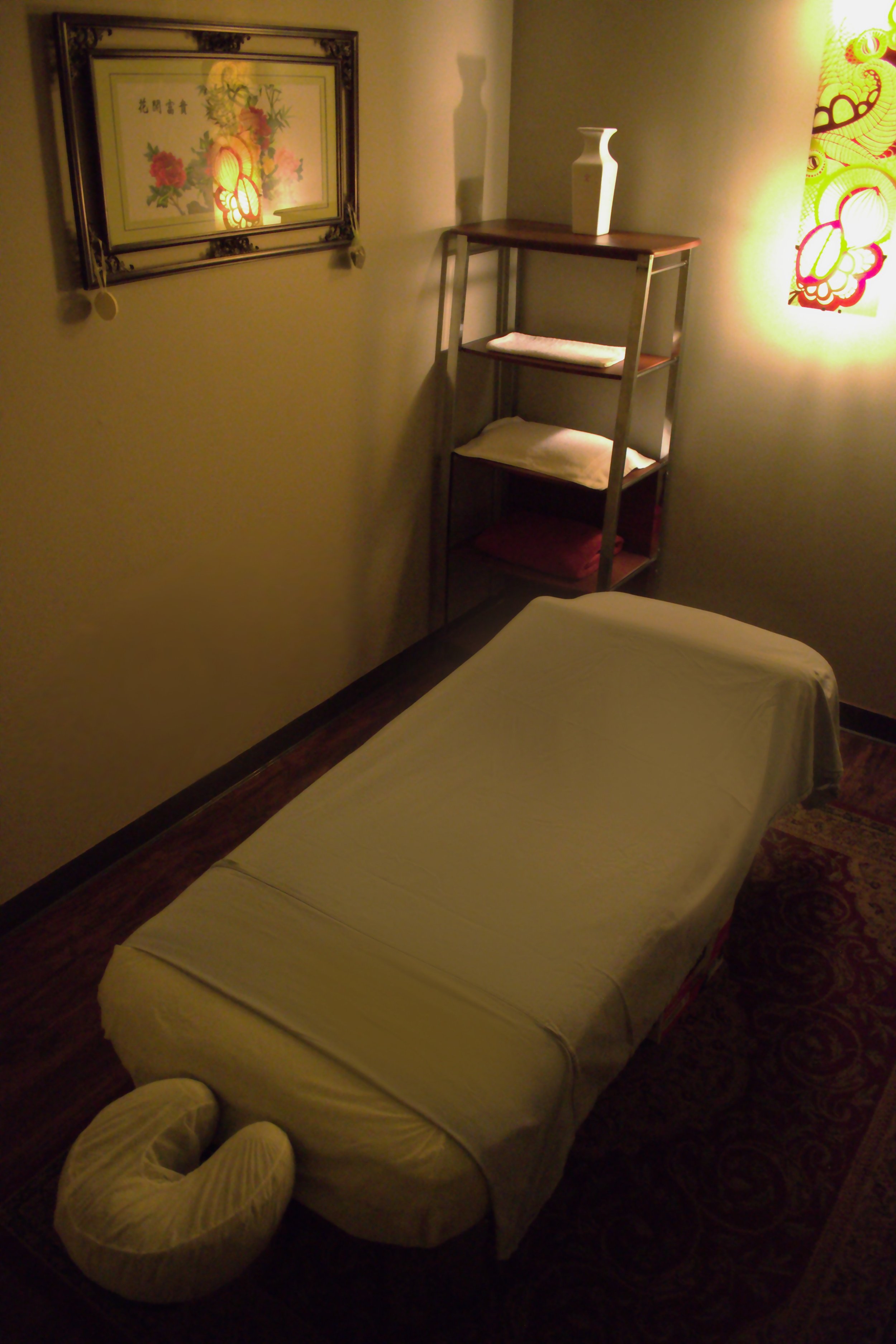 A massage table covered with a white sheet and a U-shaped pillow at the head, situated in a dimly lit room with wall art and a shelf with folded towels and vases.
