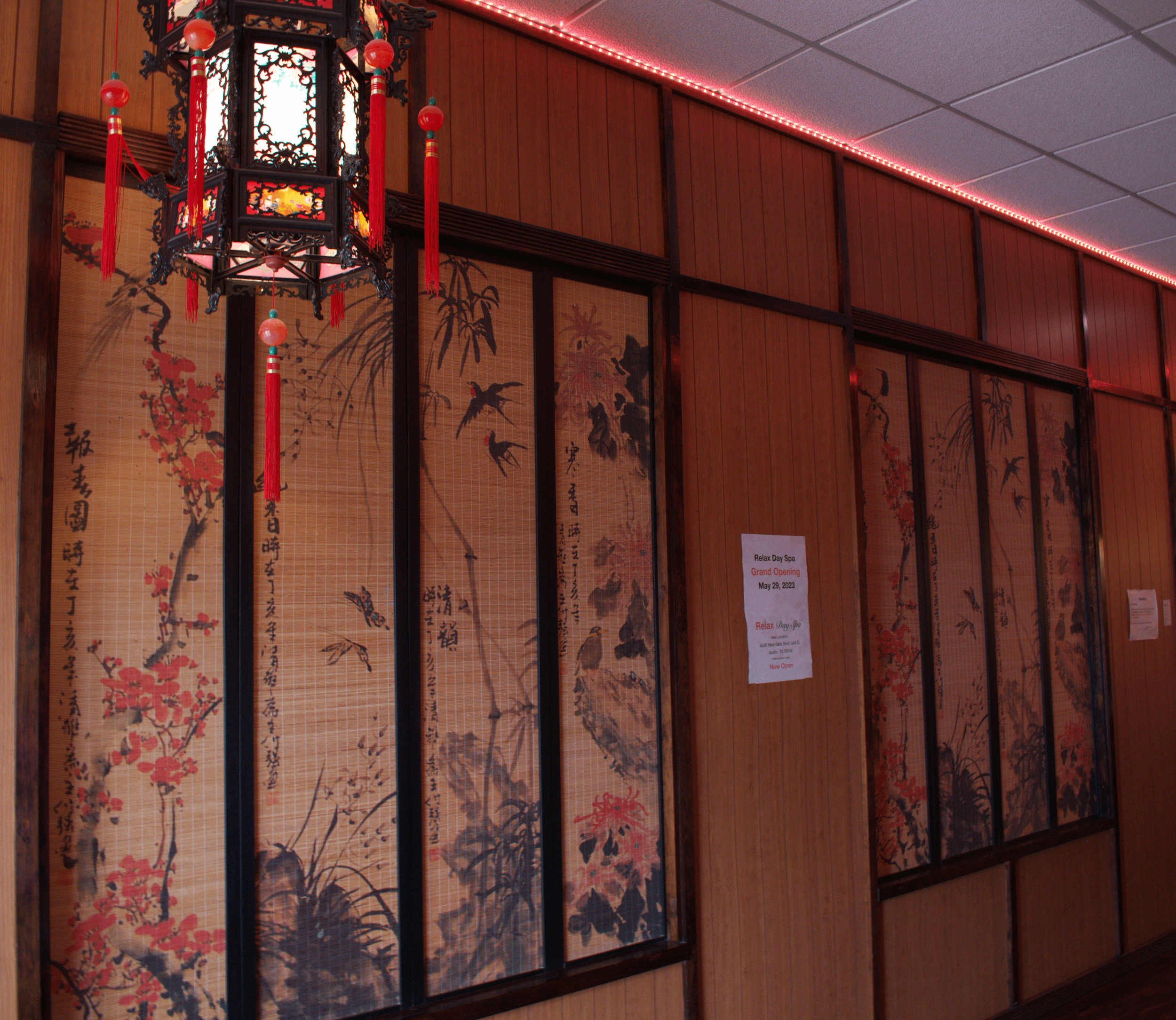 Interior of a Chinese-themed spa or restaurant with wooden wall panels, decorative paintings of birds and flowers on bamboo mats, a traditional Chinese lantern hanging from the ceiling, and a sign on the wall announcing the grand opening.