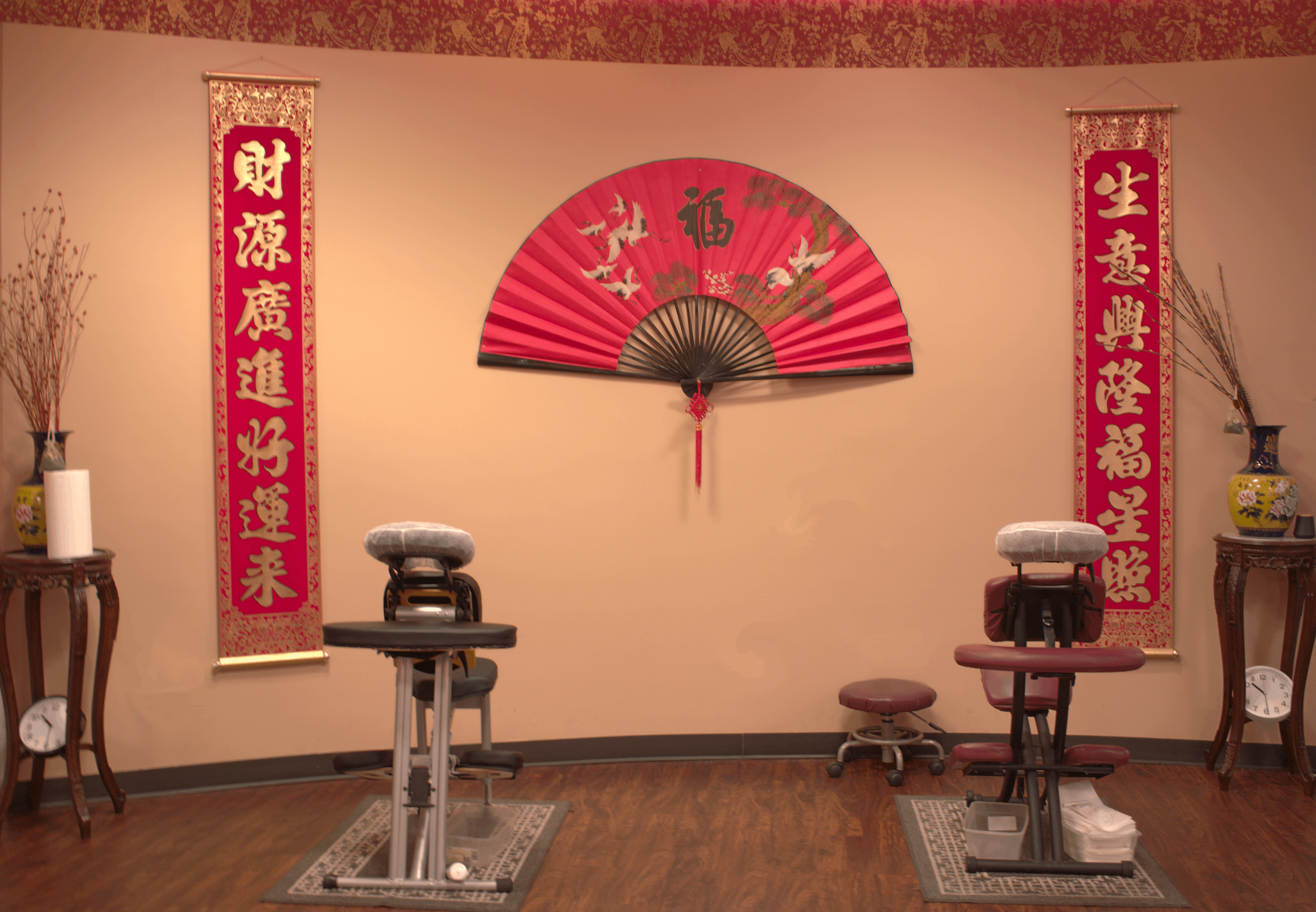 Interior of a massage or acupuncture clinic with traditional Asian decor, including two vertical red banners with gold Chinese characters on each side, a decorative red fan with cranes and the character for 'fortune' in the center, massage tables, and vases with dried branches.
