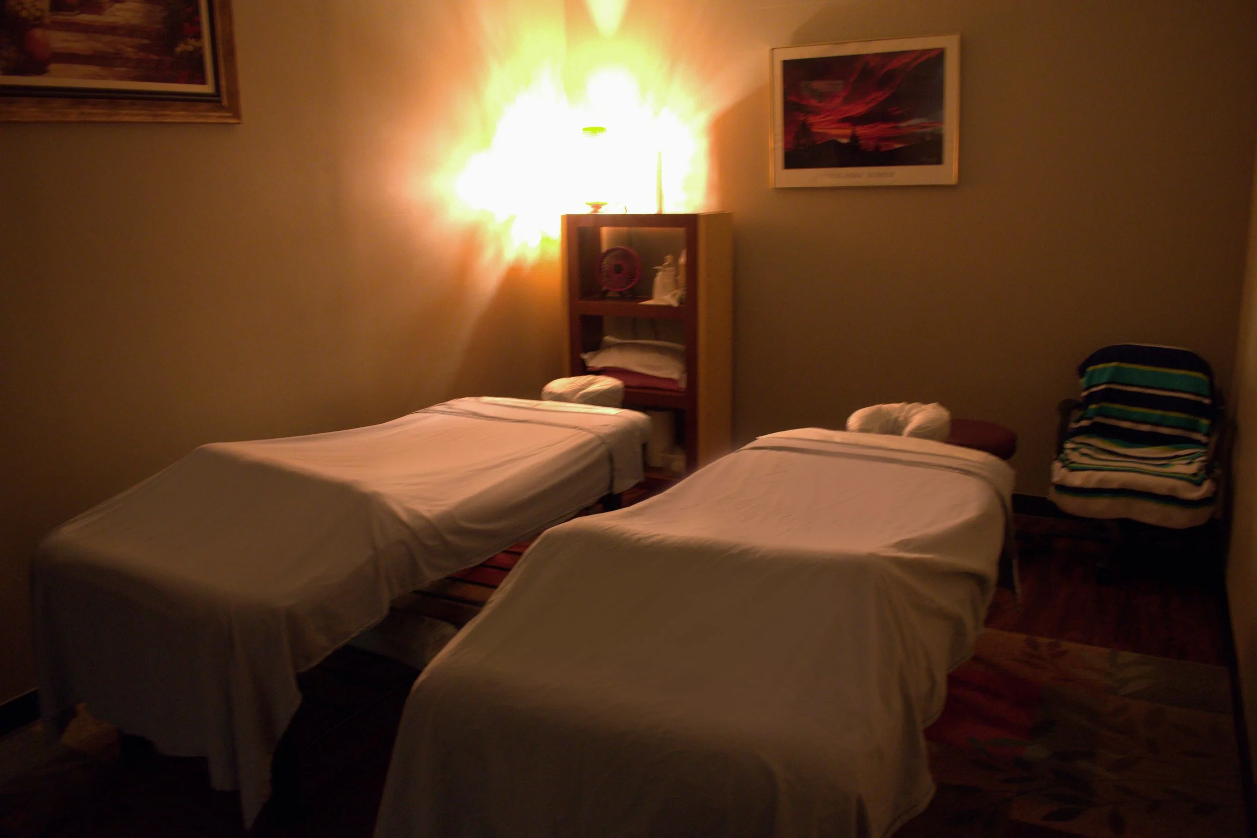 A dimly lit room with two massage tables covered in white sheets, a small bookshelf with towels and items, a lamp casting a warm glow, framed pictures on the wall, and a stack of striped towels or cloths on a chair.