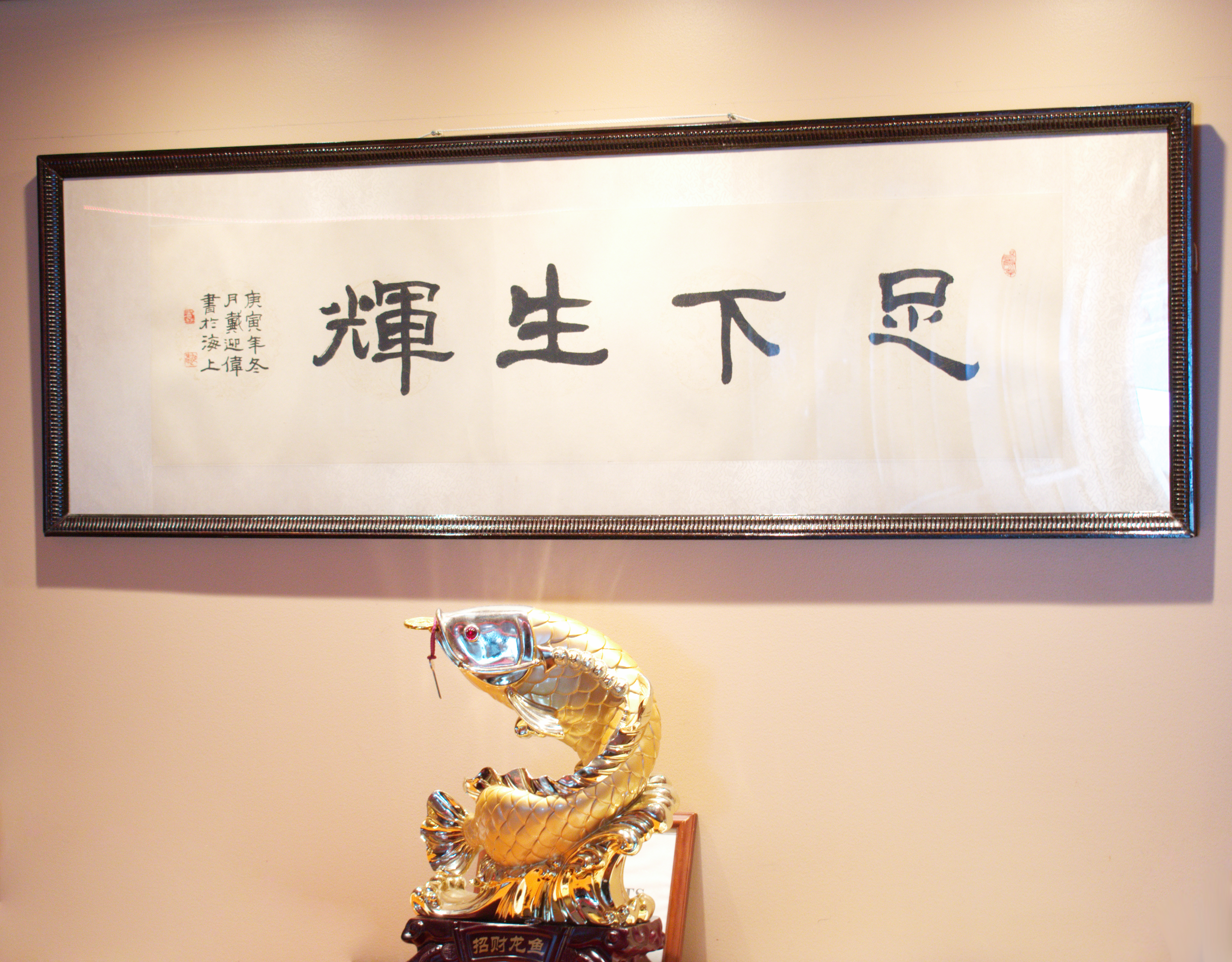 A framed piece of Chinese calligraphy artwork hanging on a beige wall, with a gold fish sculpture placed below it.