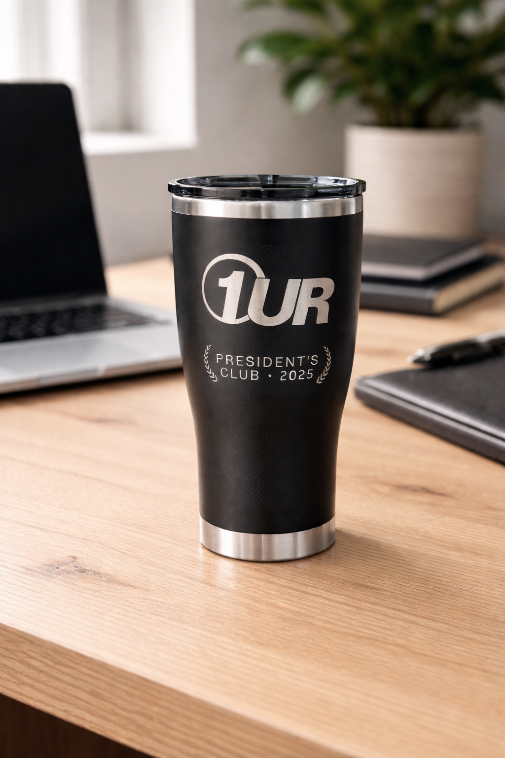 A black tumbler with silver accents and the text '1UR President's Club 2025' on a wooden desk, with a laptop, notebooks, and a plant in the background.