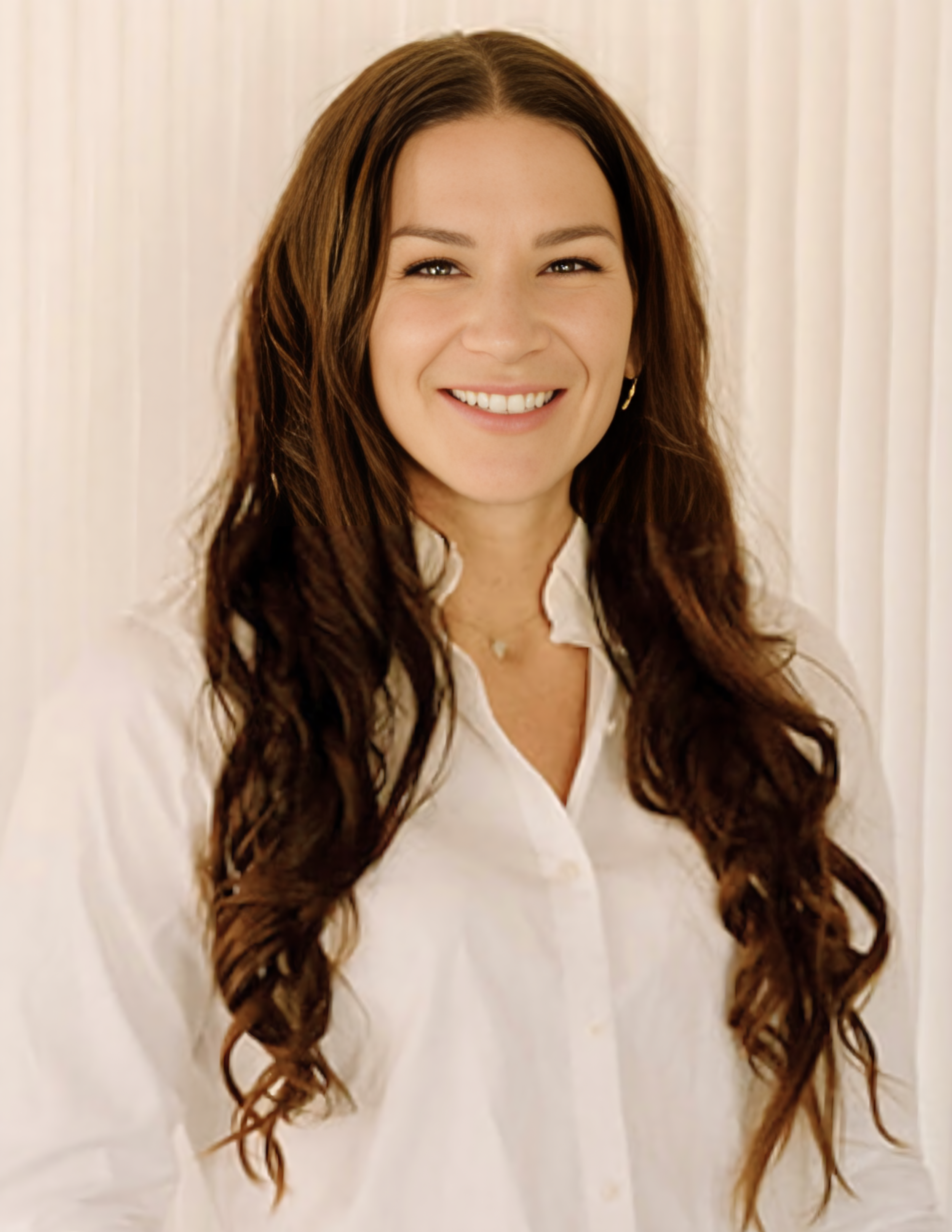 A woman with long, wavy brown hair, wearing a white shirt and smiling at the camera.
