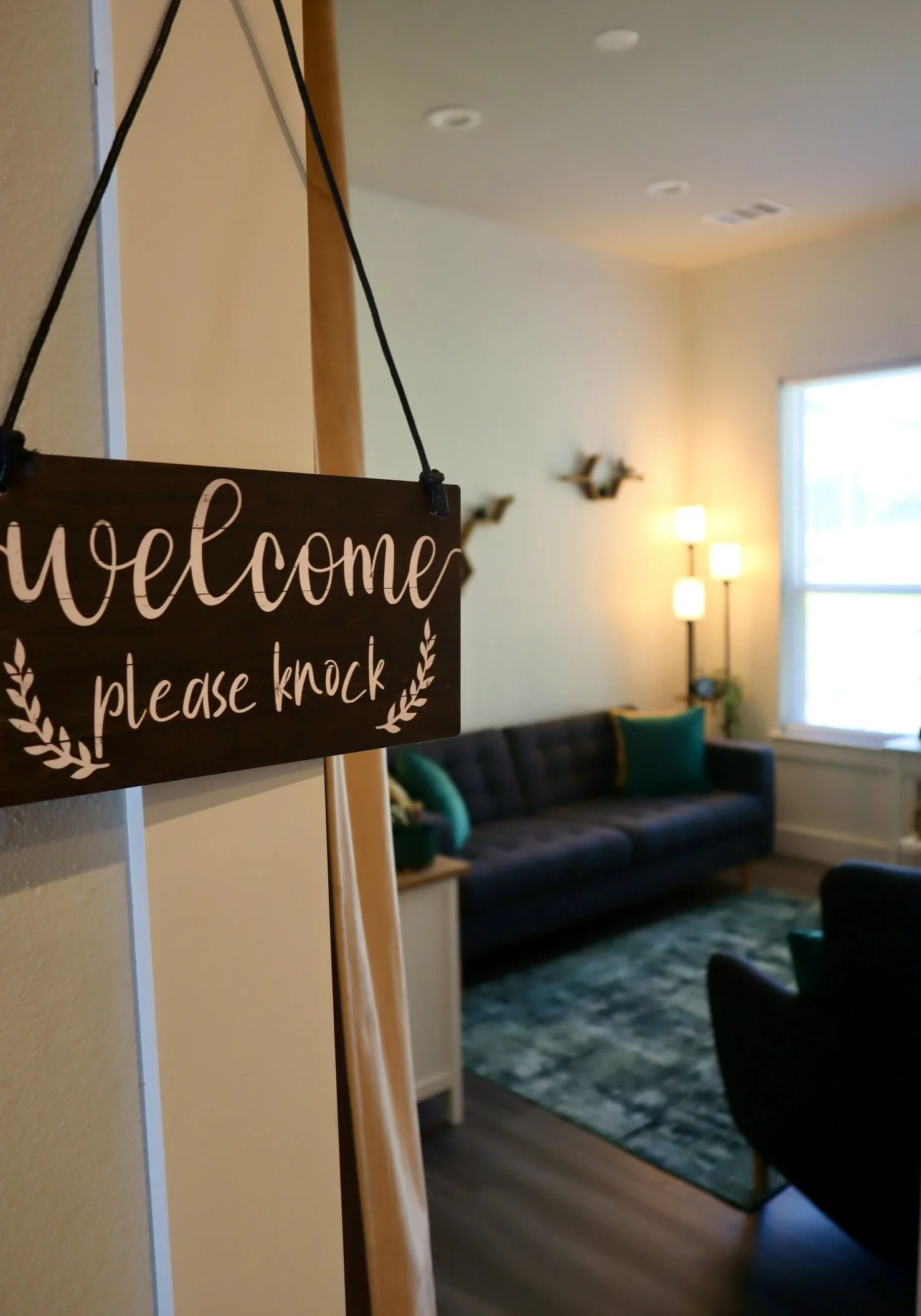 A welcome sign hanging on a doorframe with a living room in the background, featuring a couch, pillows, lamps, and a window.