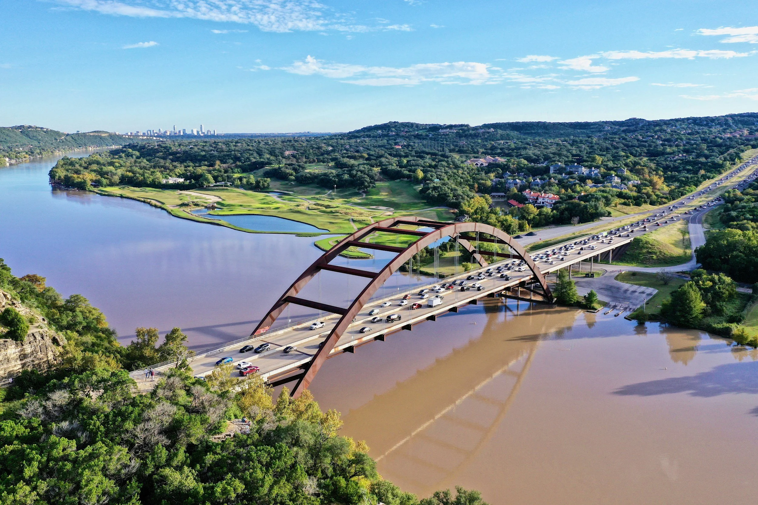 Aerial view of a bridge over a river with traffic, with lush green landscape and golf course nearby, and a city skyline in the distance.