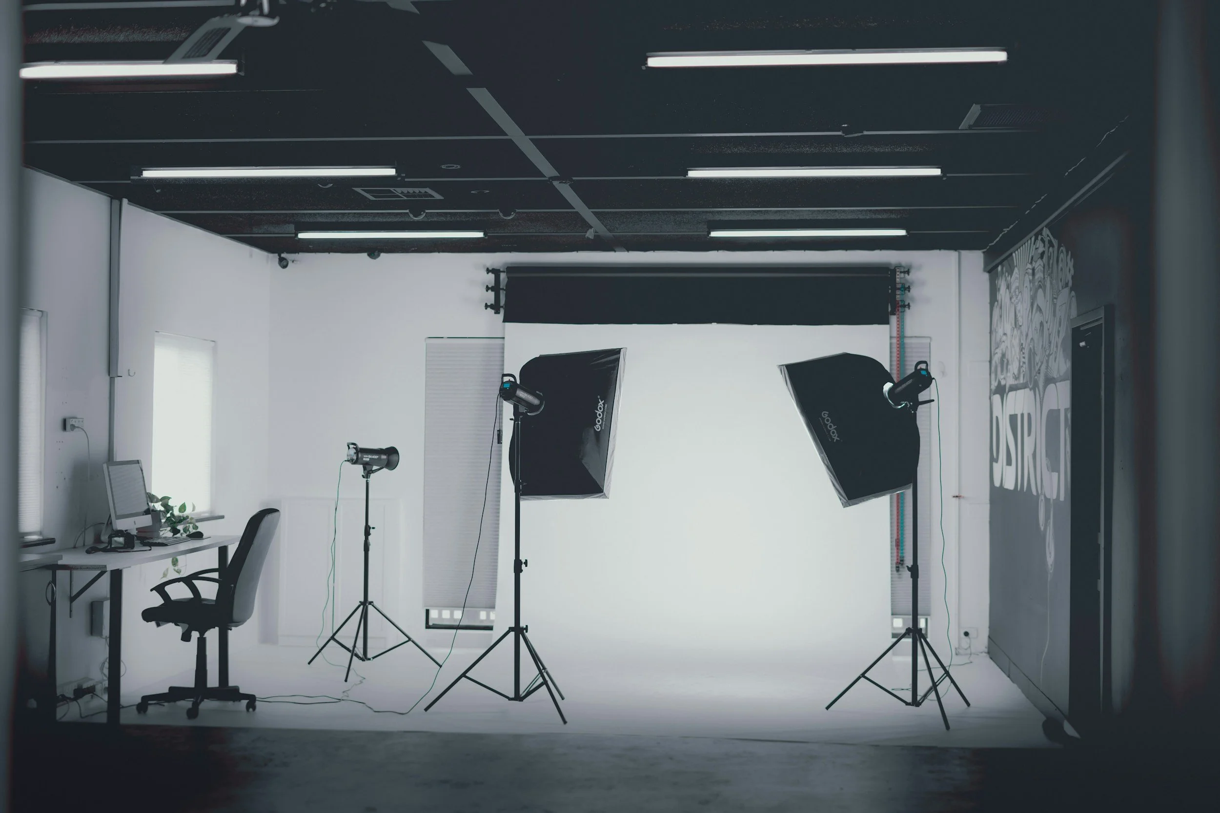 Photography studio with a white backdrop, two softbox lights on tripods, a computer on a desk, and a black office chair, setting up for a photoshoot or video production.