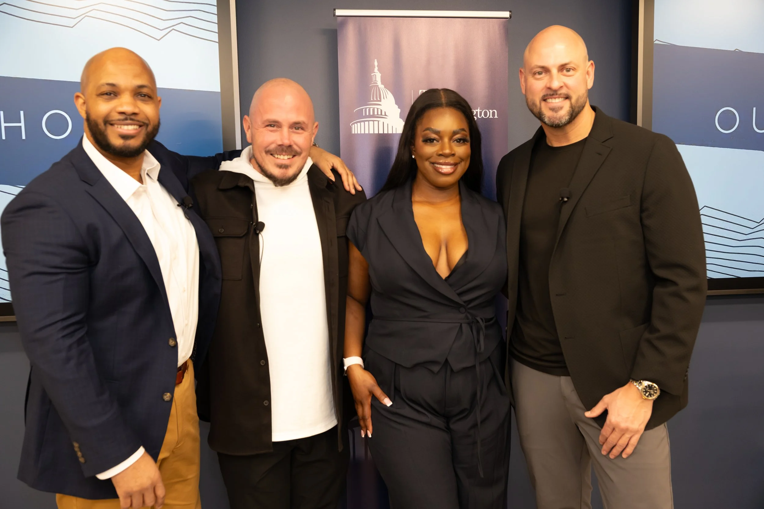Four people standing together and smiling, two men and two women, at an indoor event, with a backdrop featuring the U.S. Capitol building, part of a presentation or gathering.