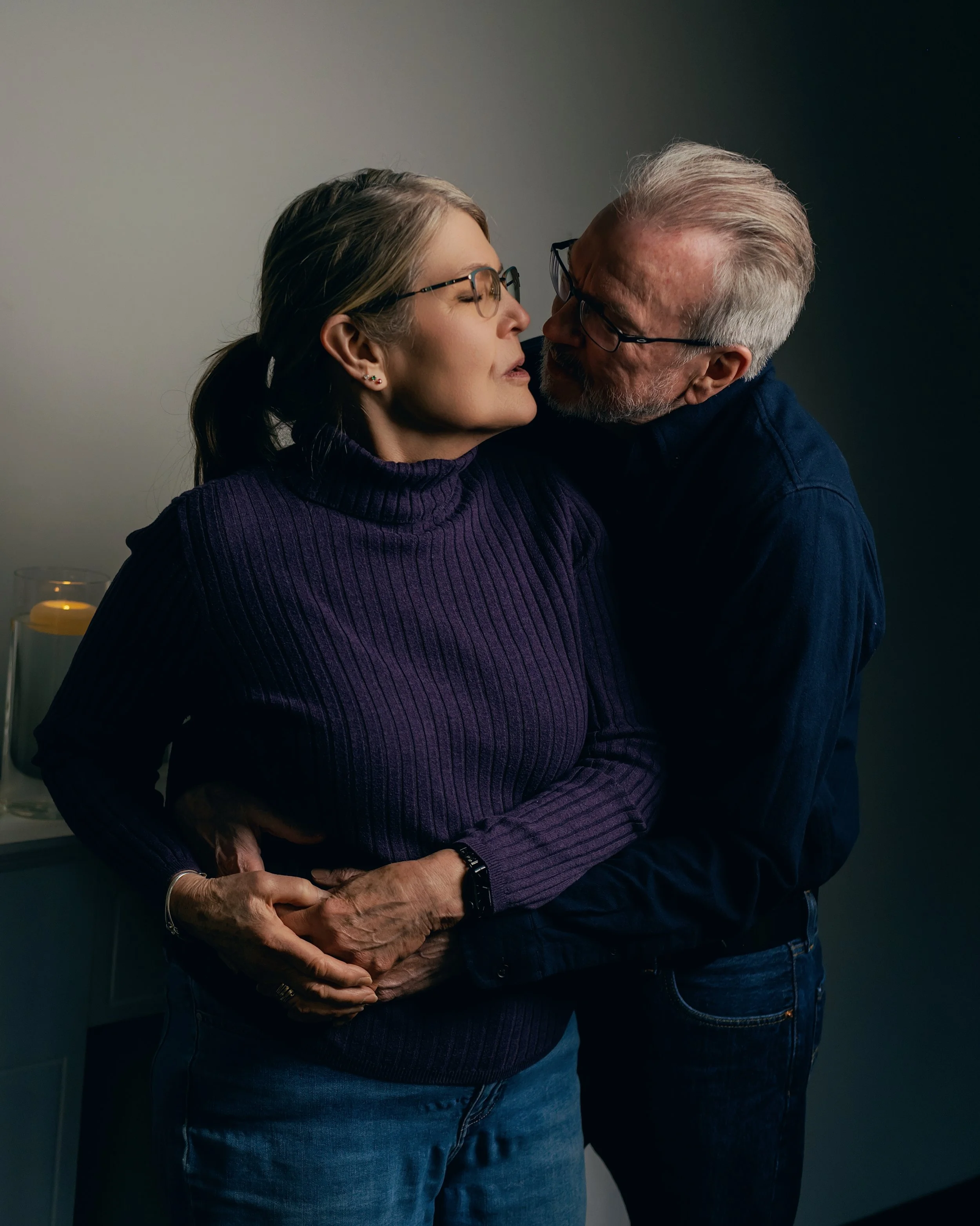 An older man and woman are close together, about to kiss, with the woman leaning slightly back and the man leaning forward. The woman has long hair tied back, glasses, and is wearing a purple turtleneck sweater. The man has gray hair, glasses, and is