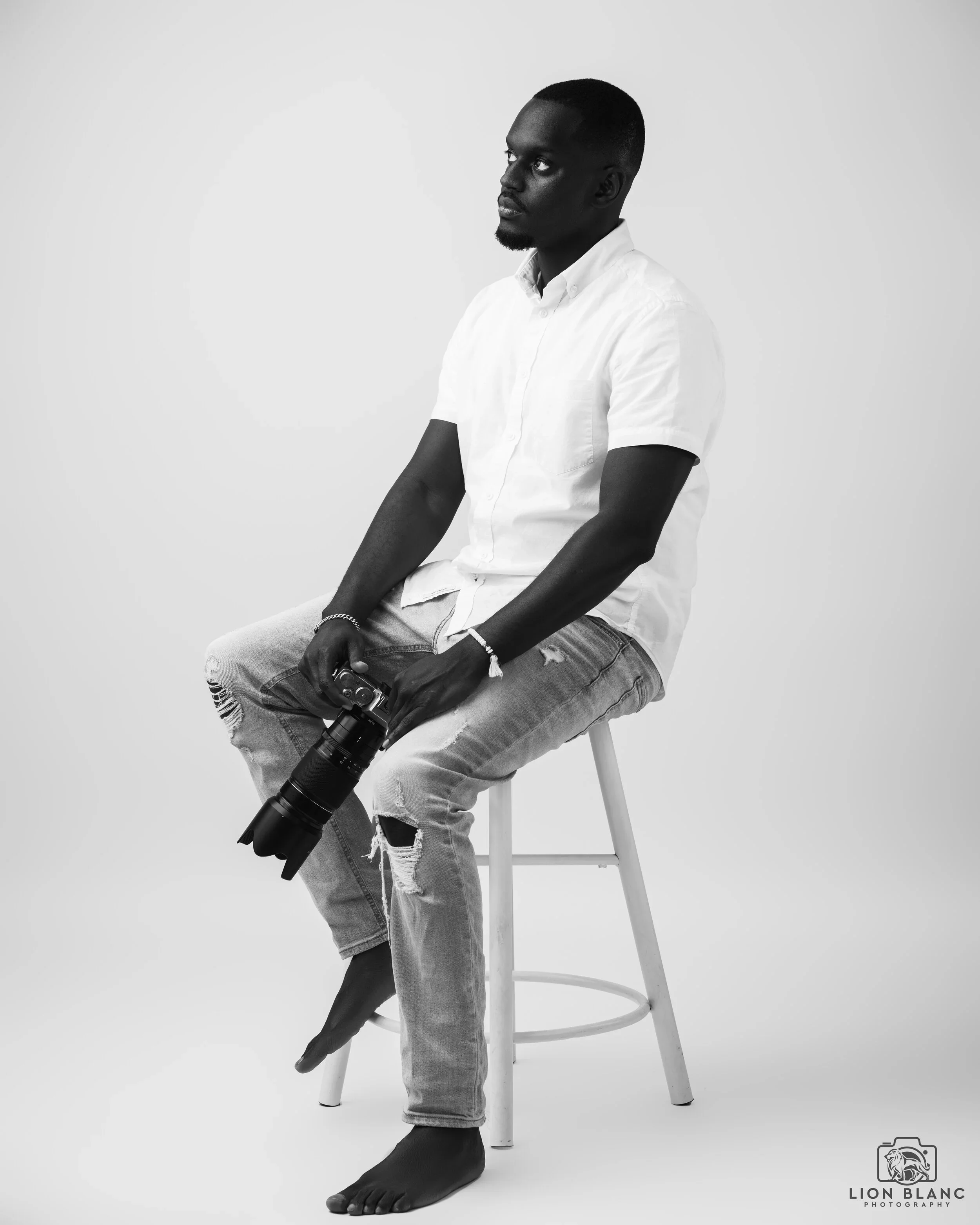 Black and white photo of a man with dark skin, sitting on a stool, holding a camera with a lens. He is barefoot, wearing a white short-sleeved shirt and ripped jeans, with a focused expression.