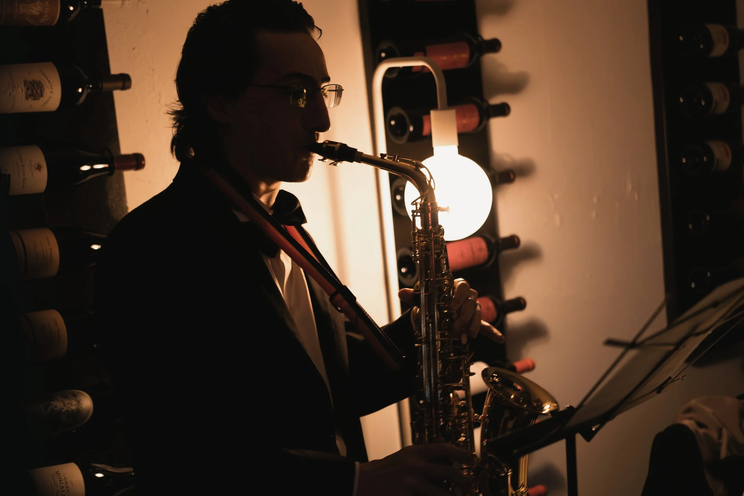 A person playing a saxophone in a dimly lit room, with wine bottles on shelves and a round light fixture in the background.