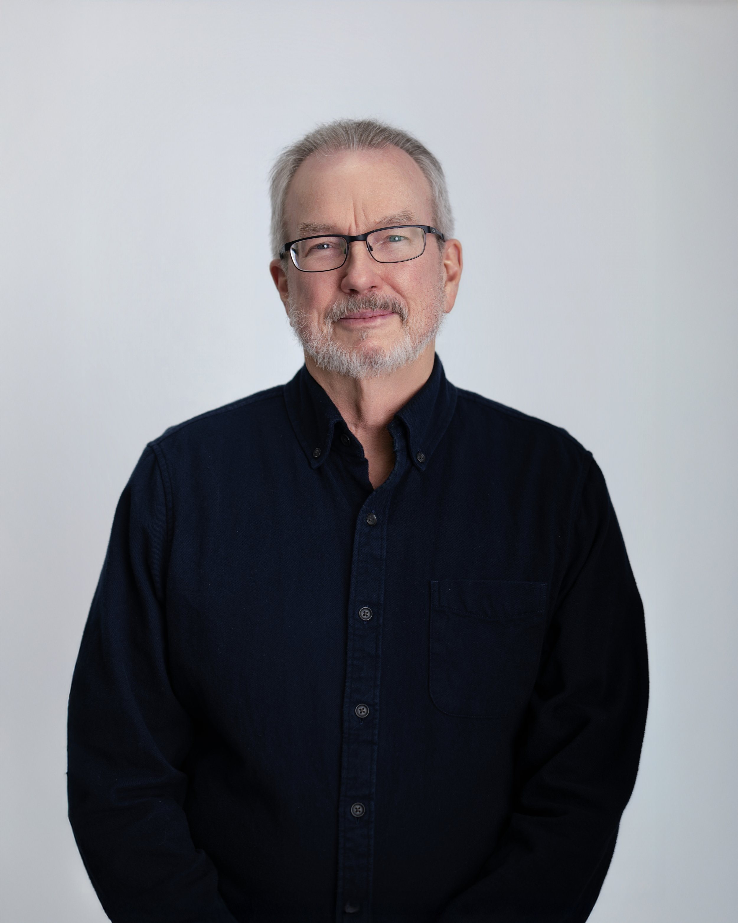 A middle-aged man with glasses, grey hair, and a beard, wearing a black button-up shirt, stands against a plain white background.