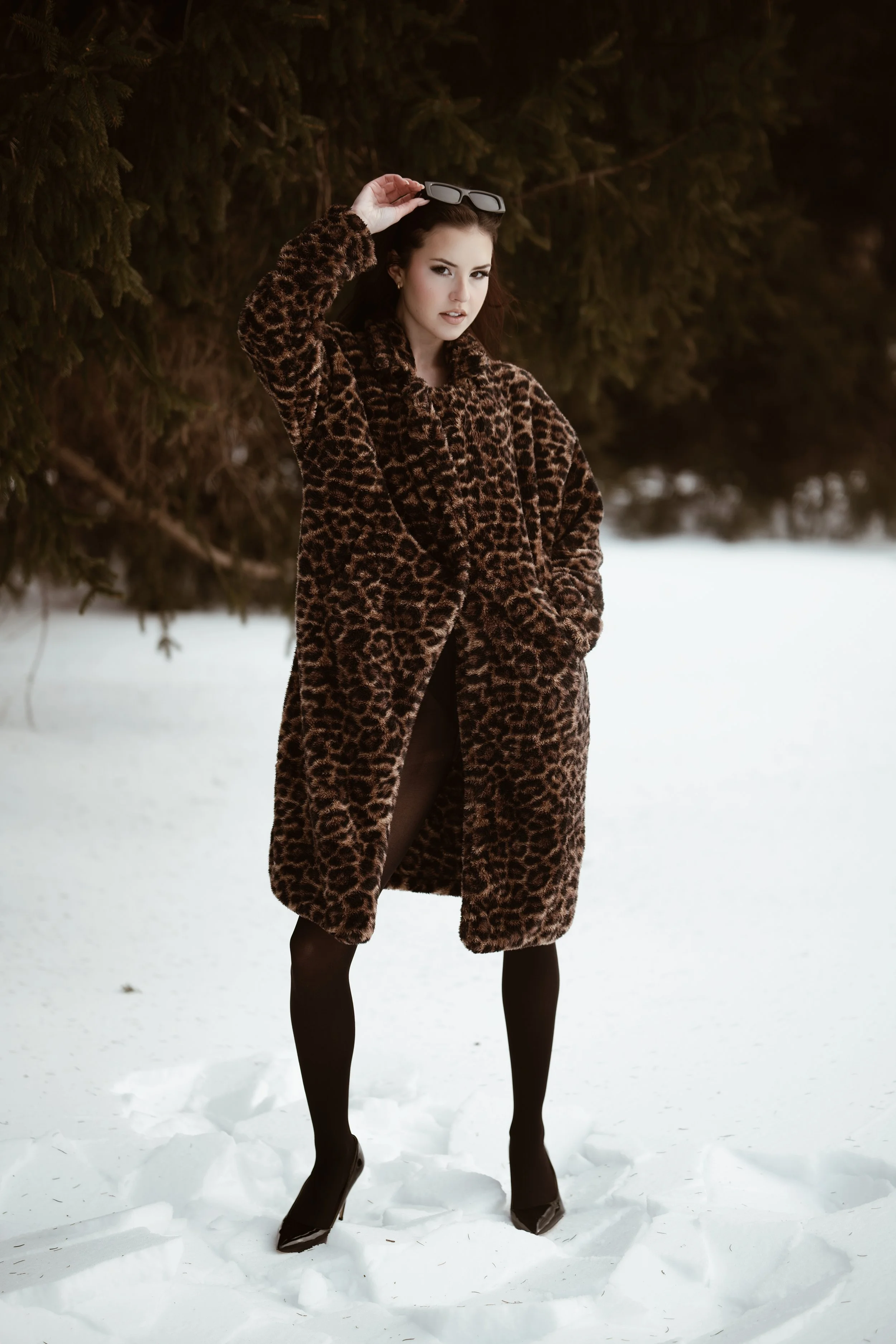 A woman standing outdoors in snow, wearing a brown leopard print fur coat, black tights, black high heels, and holding sunglasses above her head, with trees in the background.