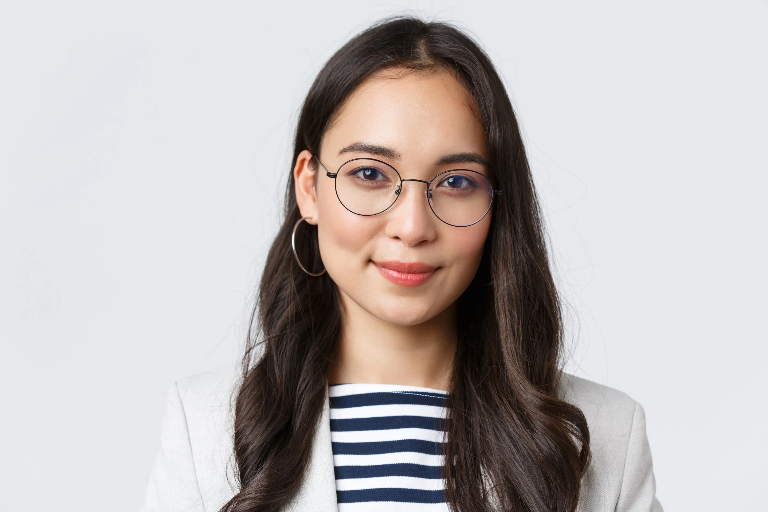 Portrait of a young woman with long dark hair, wearing glasses, hoop earrings, a striped shirt, and a light blazer, smiling against a plain white background.