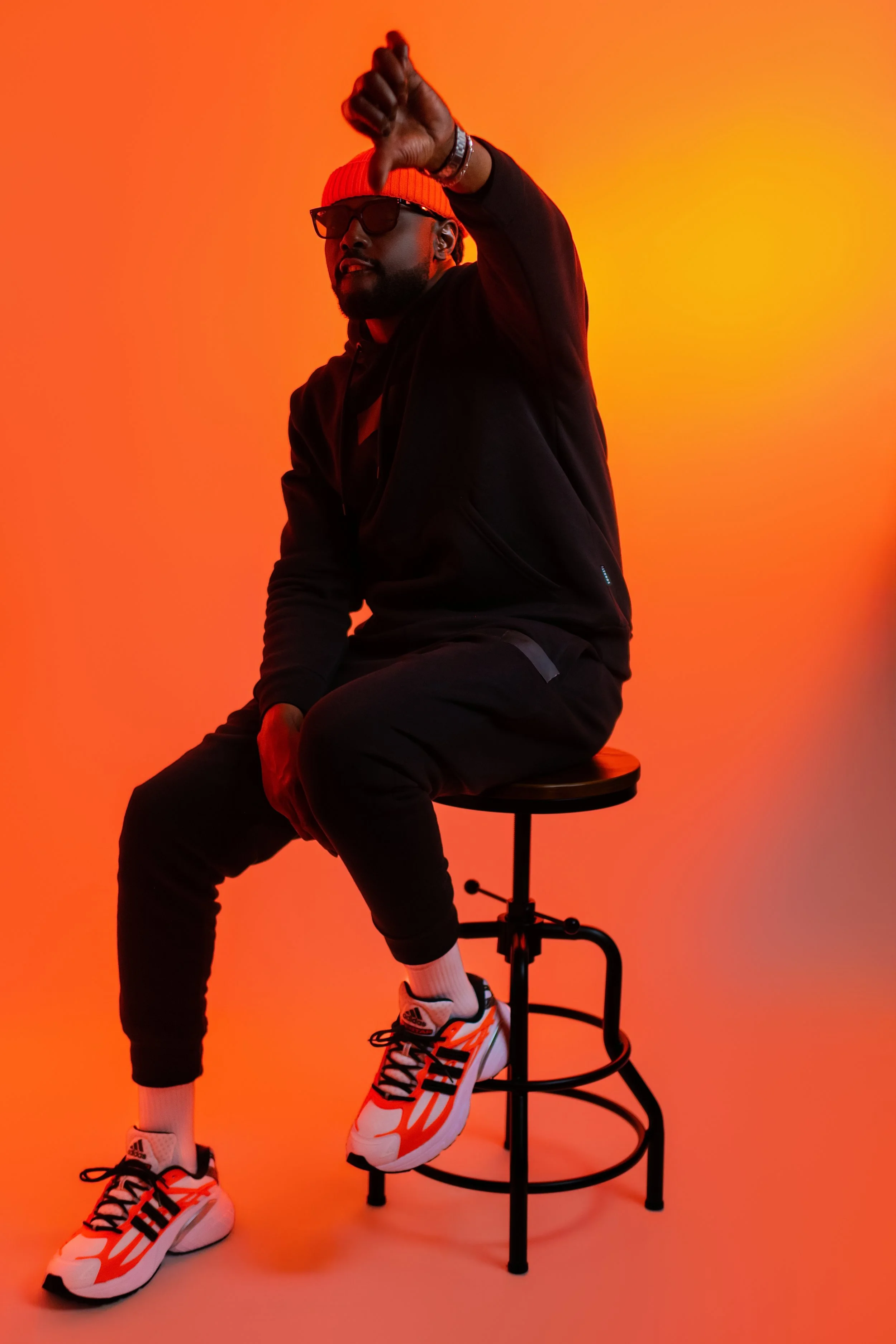 A man sitting on a stool, wearing sunglasses, a beanie, athletic shoes, and a black hoodie, against an orange gradient background, making a gesture with his hand.