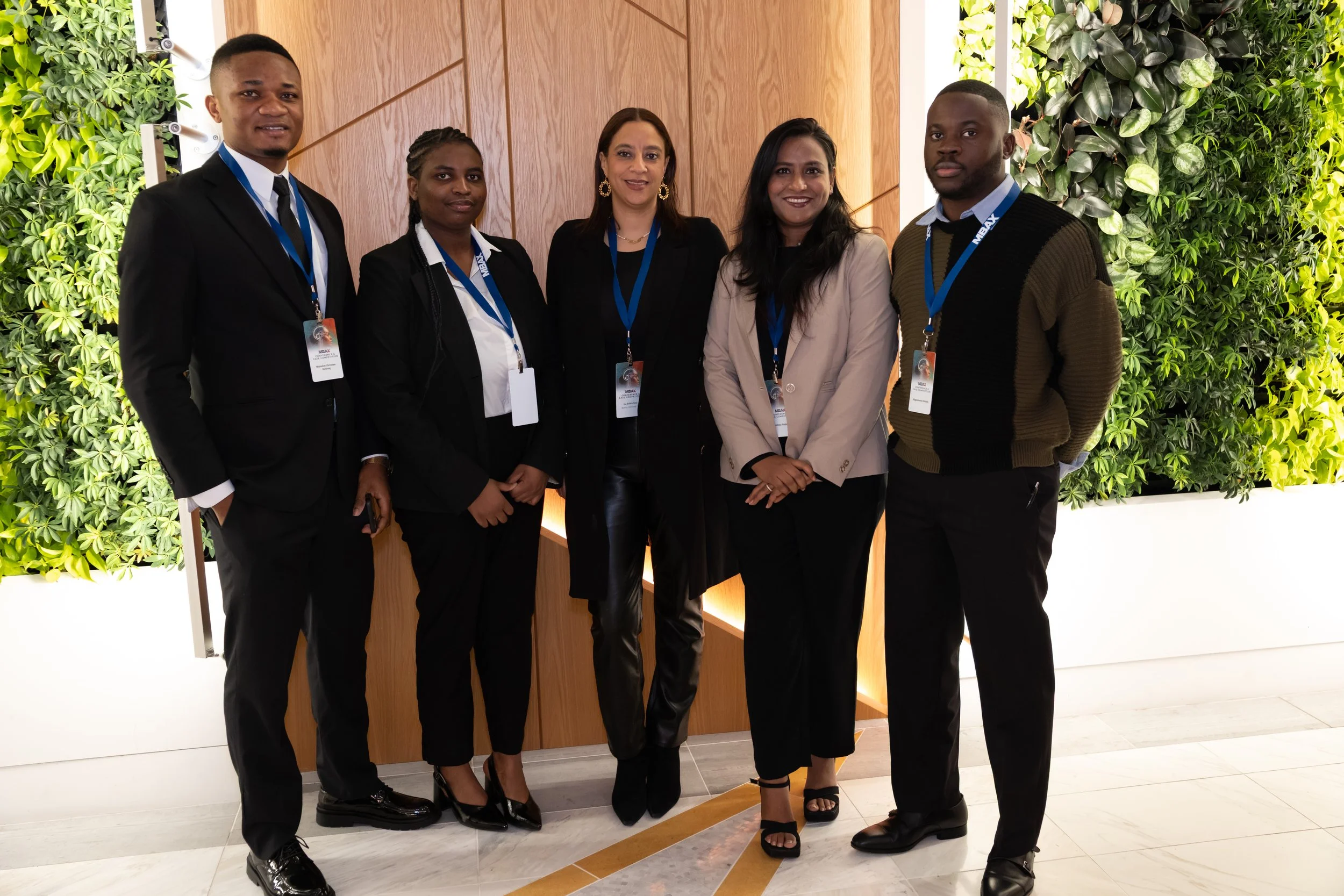 Group of five professional individuals standing together indoors at a conference or event, wearing formal business attire with name badges and lanyards, with a decorative green wall and wooden panel background.