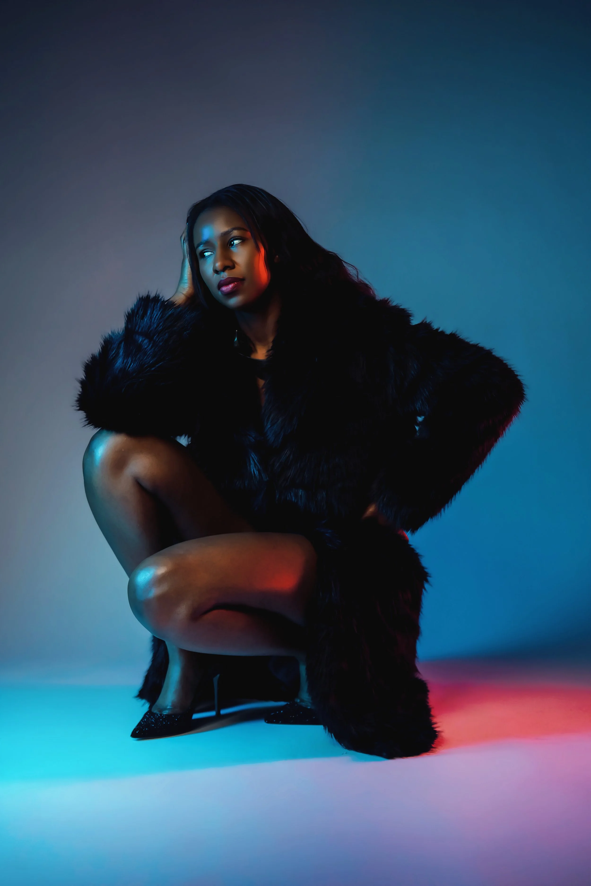 A woman squatting in a studio with colorful lighting, wearing a black fur coat and black high heels.