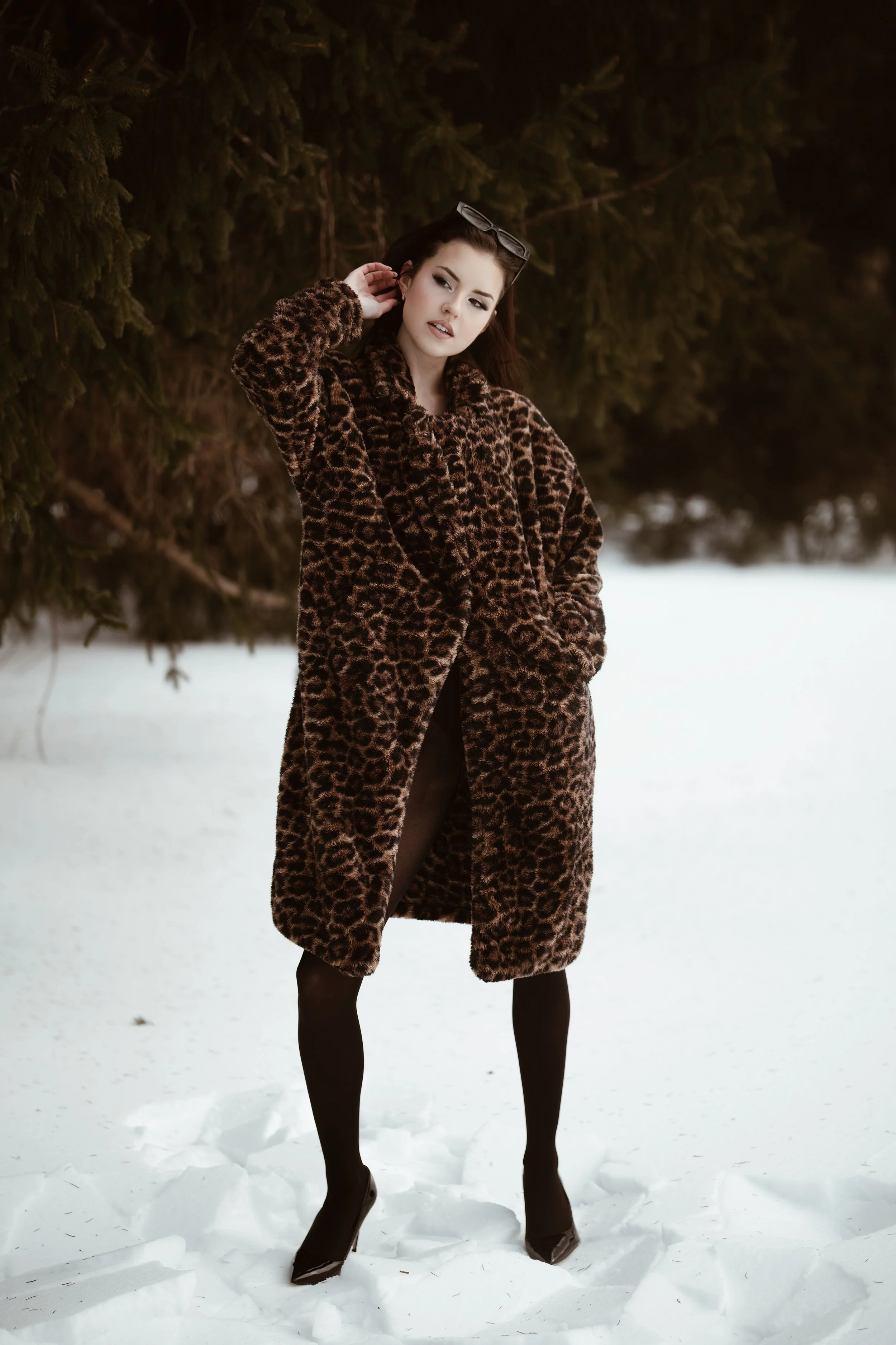 A woman wearing a leopard print coat, black stockings, and high heels stands in a snowy landscape with evergreen trees in the background.