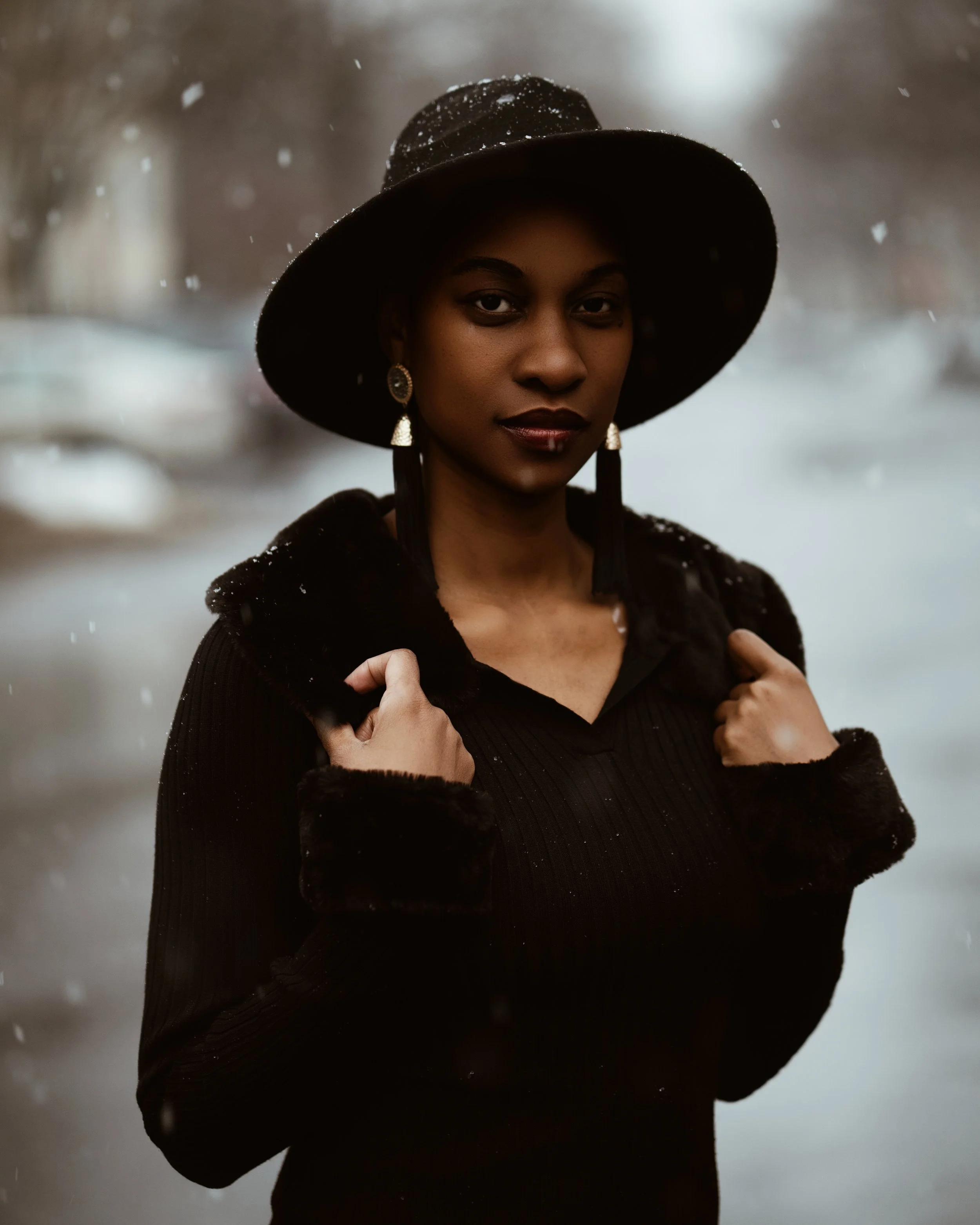 A woman in a black wide-brimmed hat and a black coat stands outdoors in a snowy scene.