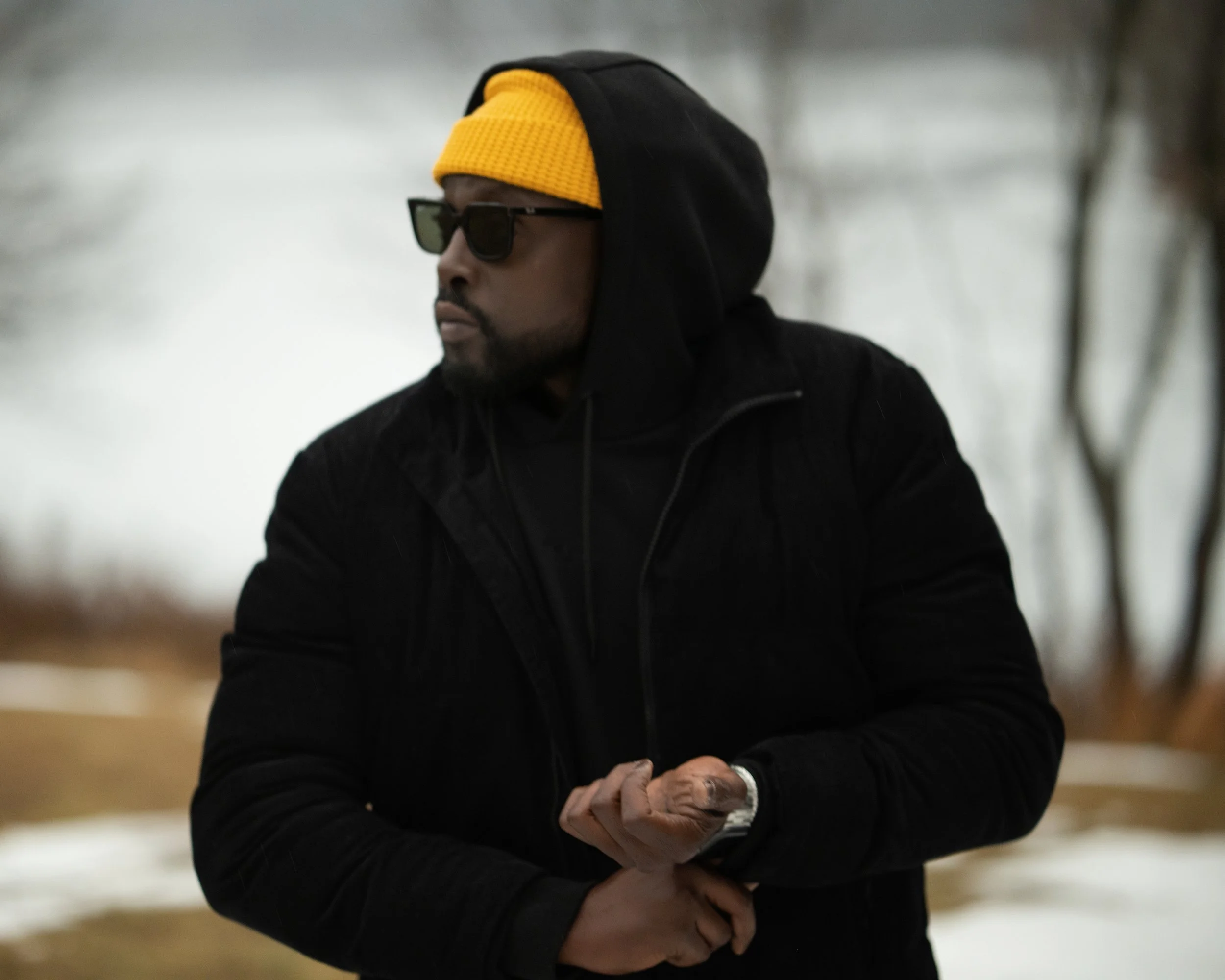 A man wearing a black hoodie, yellow hat, sunglasses, and a black jacket, standing outdoors in a wintery setting with snow and leafless trees in the background.