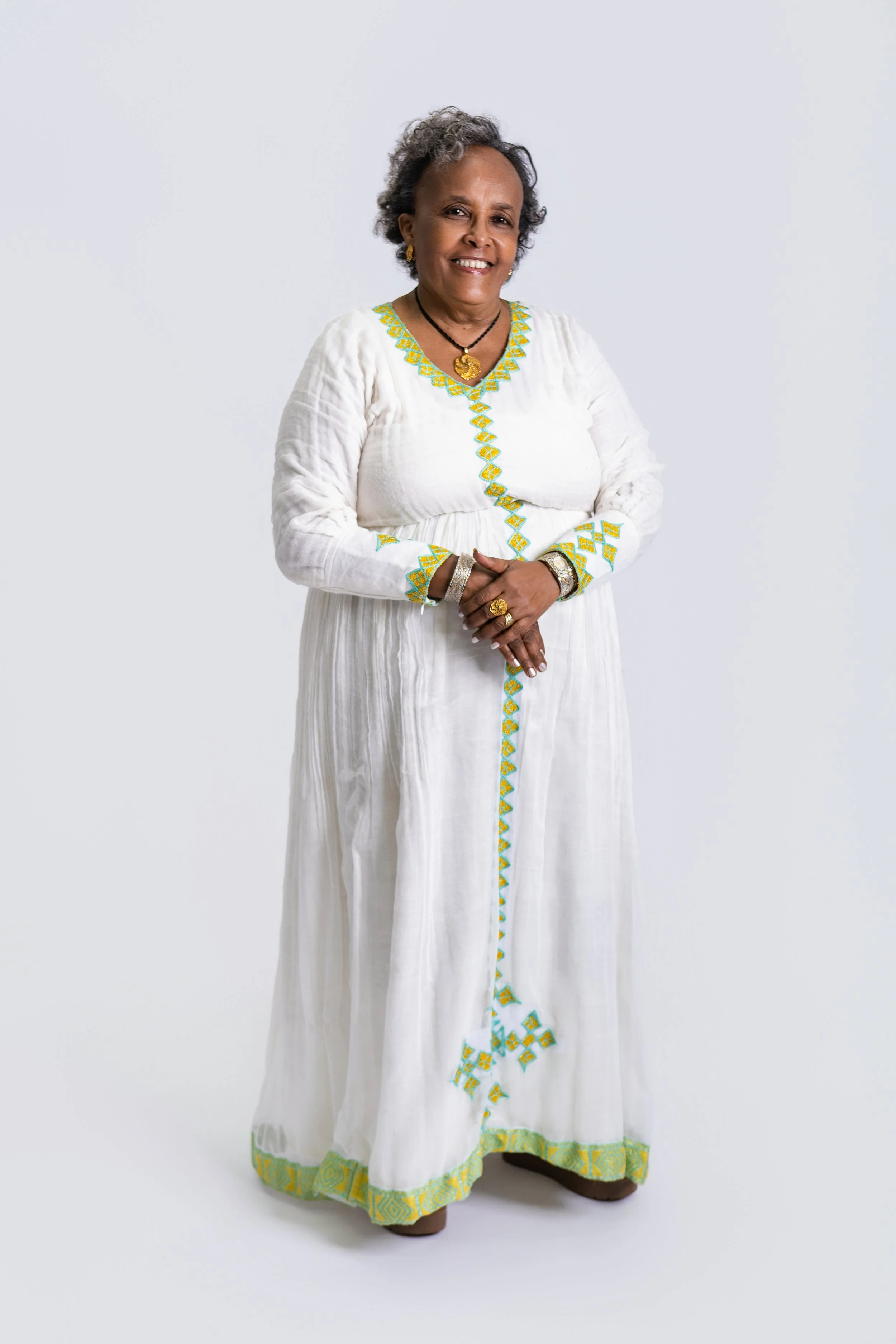 A smiling older woman with short gray hair, wearing a long white dress with yellow and green embroidery, black jewelry, and standing against a plain white background.