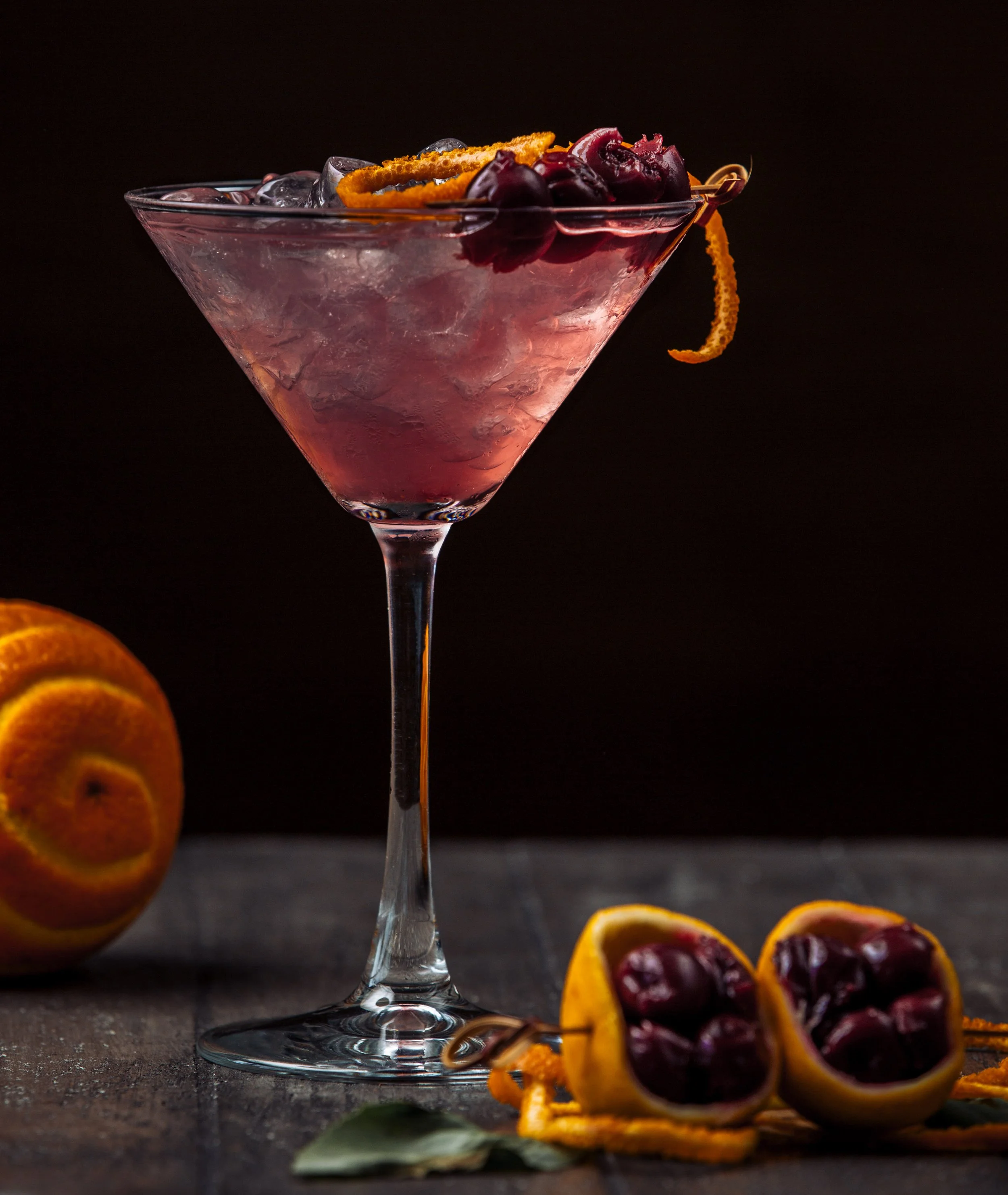 A pink cocktail in a martini glass garnished with cherries, orange zest, and a twist of orange peel, with additional cherries and orange peel on a dark wooden surface.