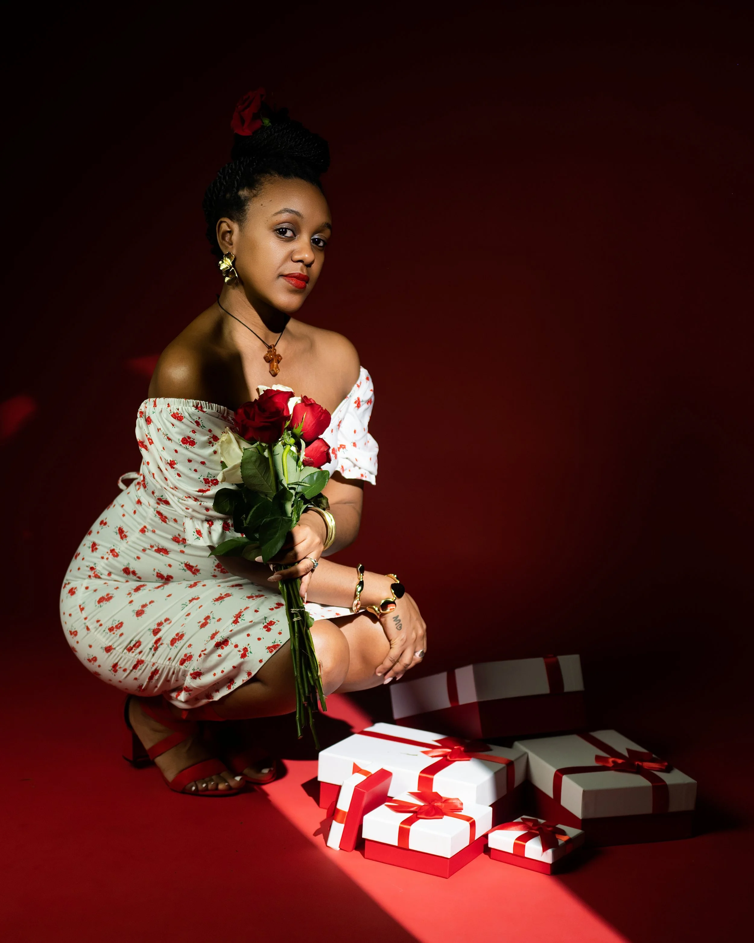 A woman in a white dress with red floral patterns, holding a bouquet of red roses, crouching beside a stack of white gift boxes with red ribbons, against a dark red background.