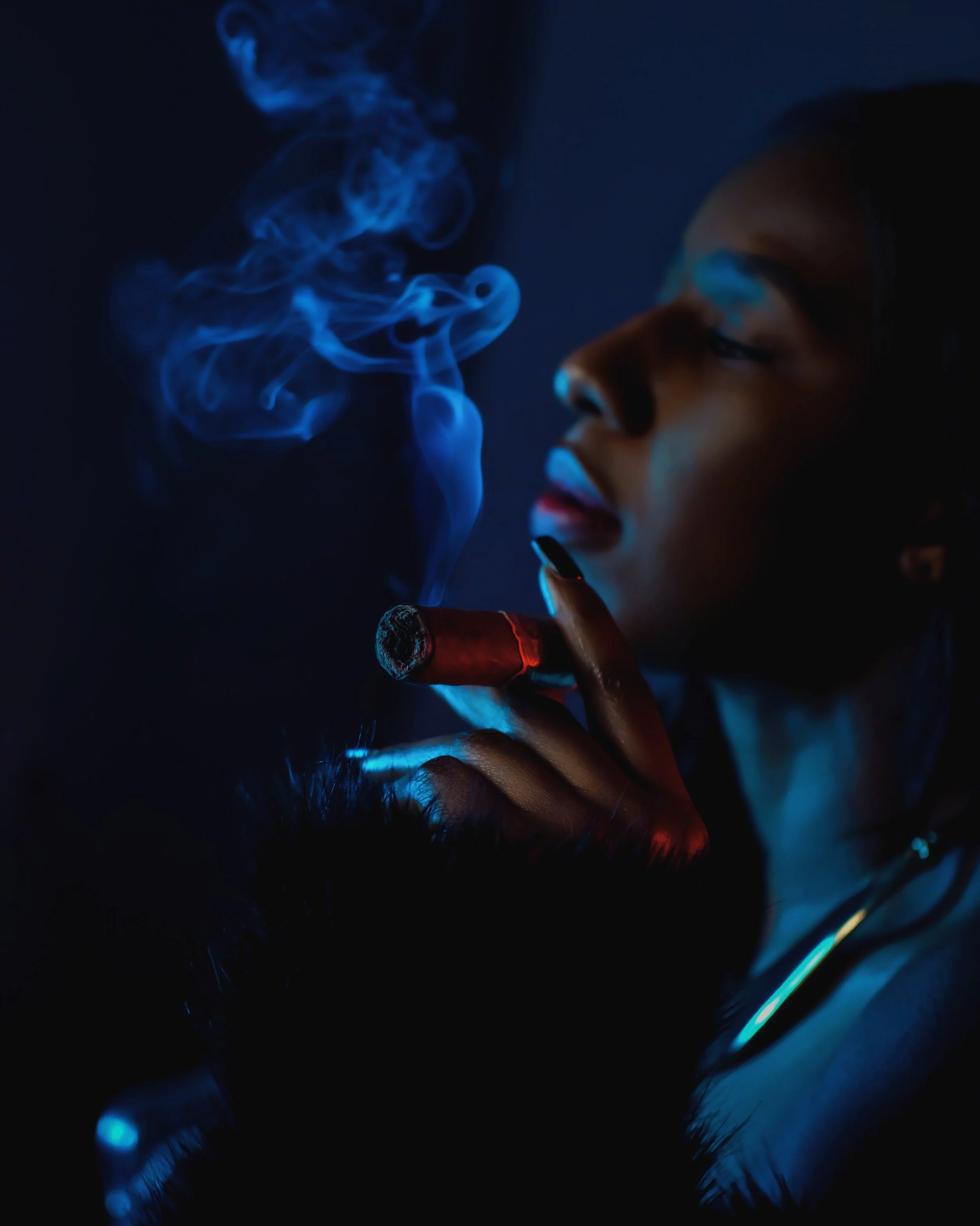 A woman with dark hair and makeup, smoking a cigarillo, with blue smoke swirling around her face in a dark setting.