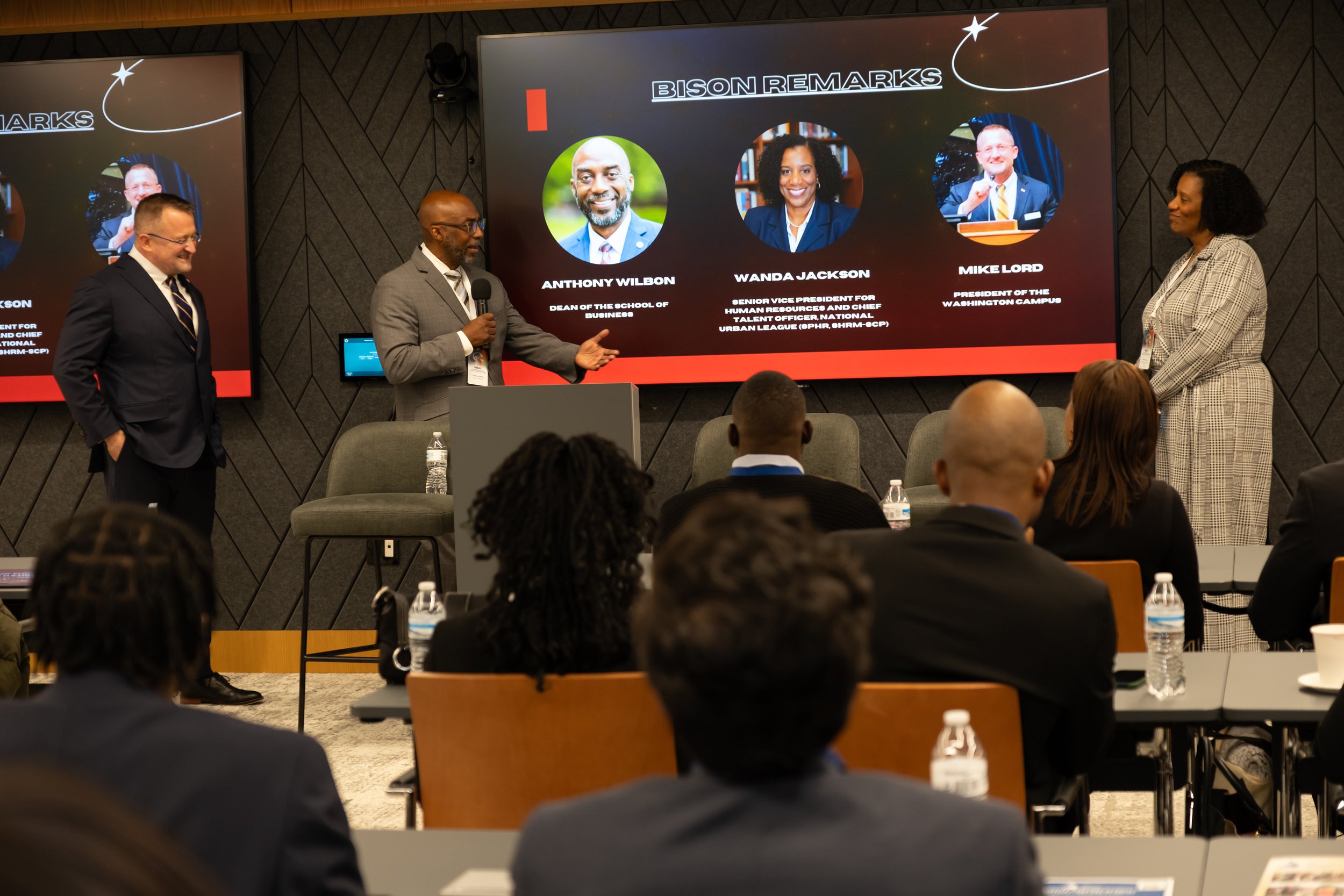 A professional conference with four speakers on stage. Three men and one woman are standing, with one man holding a microphone. The large screen behind them displays photos and titles, including Anthony Wilbon, Wanda Jackson, and Mike Lord. The audience is seated at tables with water bottles, listening to the presentation.