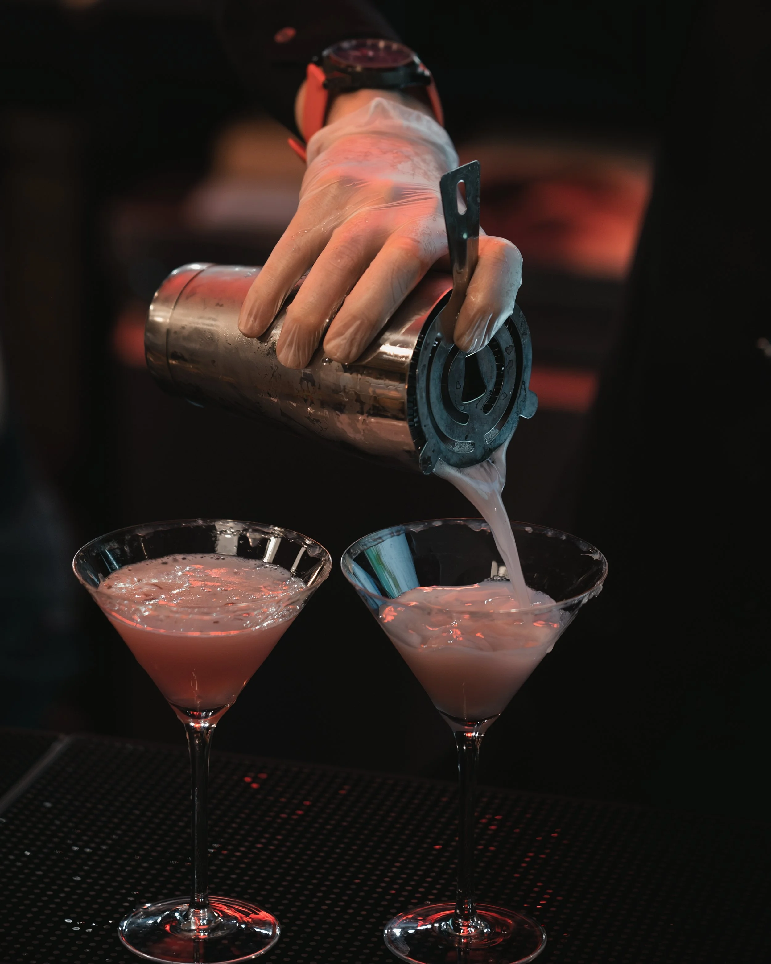 A bartender wearing gloves pours a pink cocktail from a shaker into two martini glasses on a bar counter.