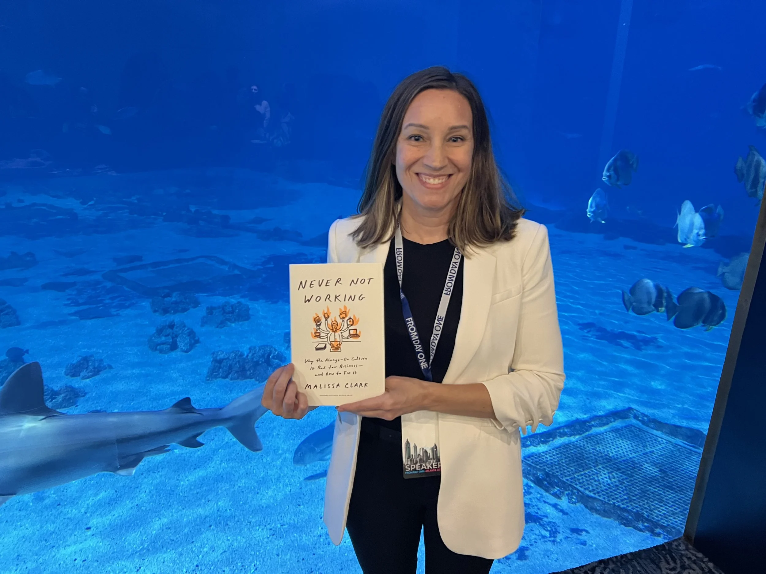 A woman smiling and holding a book titled 'Never Not Working' in front of an aquarium with sharks and fish.