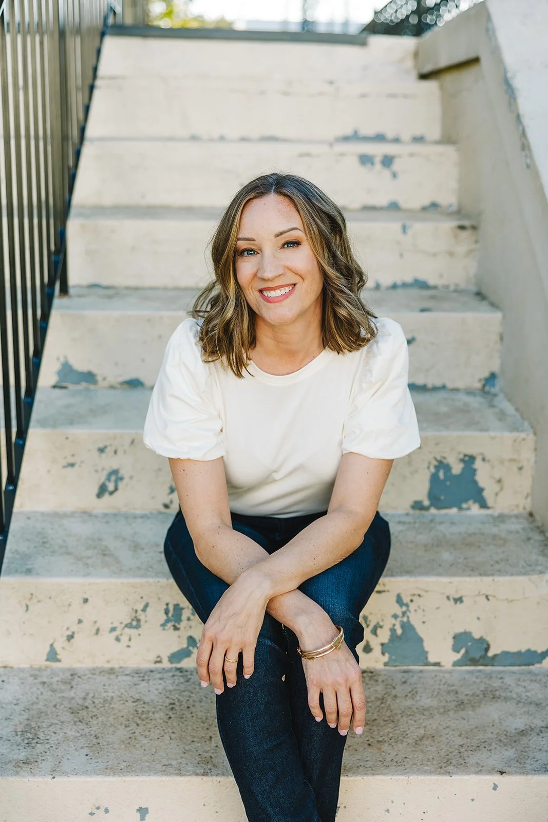 A smiling woman with shoulder-length wavy brown hair, wearing a white short-sleeve shirt and dark jeans, sitting on concrete stairs with chipped paint and a metal railing on the side, outdoors.