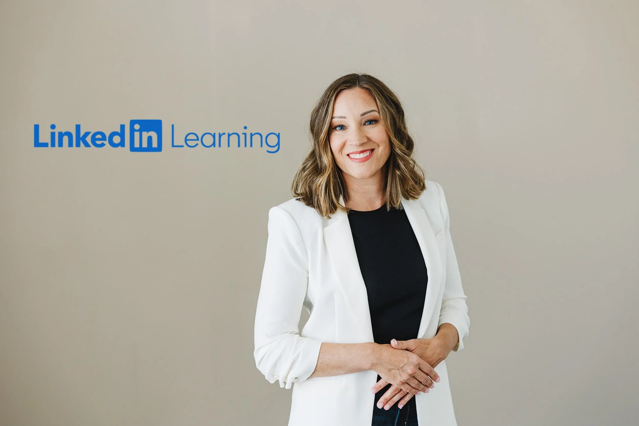 A woman with shoulder-length brown hair, smiling, wearing a white blazer over a black top, standing in front of a plain beige wall with the blue LinkedIn Learning logo.