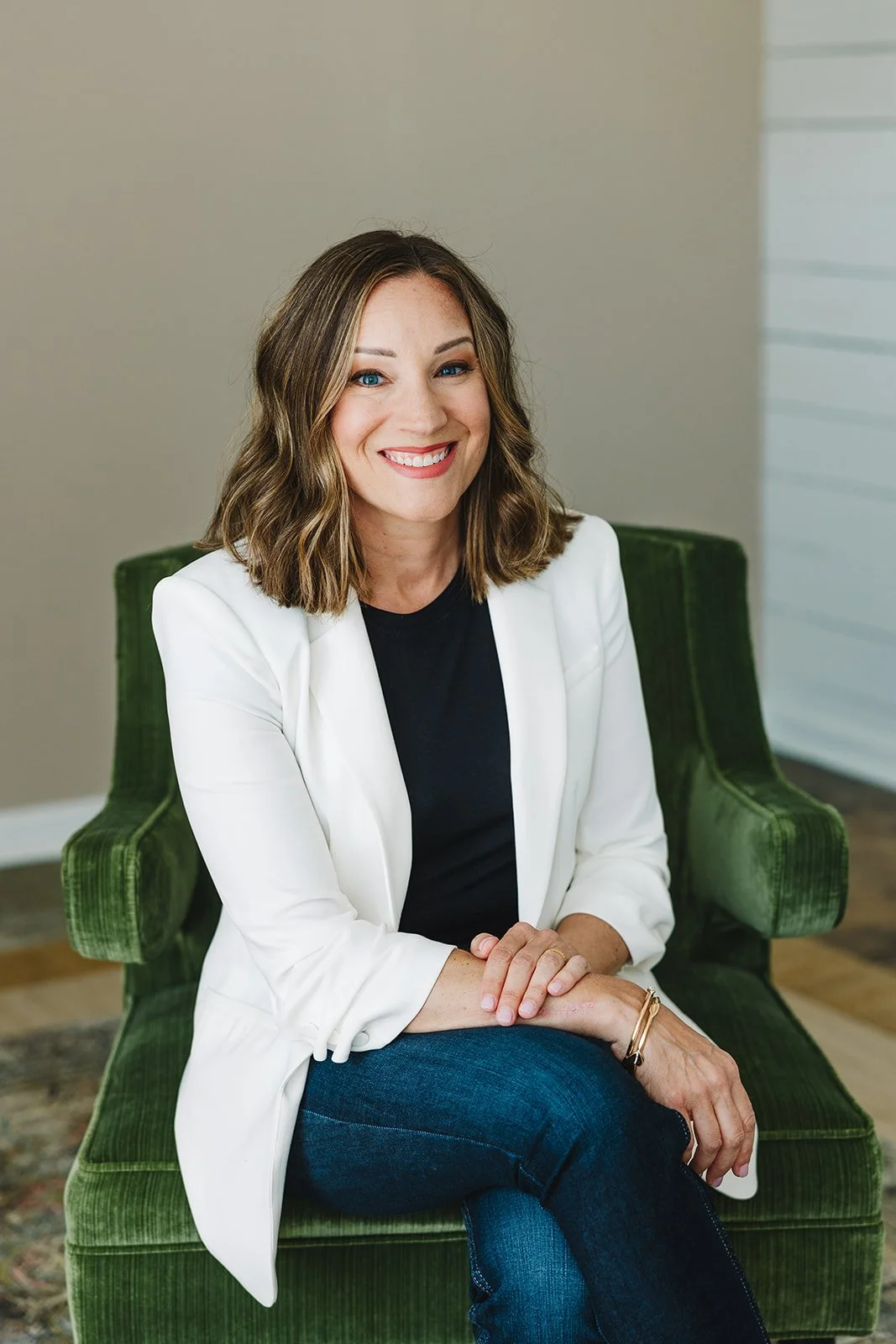 A woman with shoulder-length wavy brown hair, blue eyes, and fair skin, smiling and sitting on a green velvet chair, wearing a white blazer over a black shirt and blue jeans in an indoor setting.