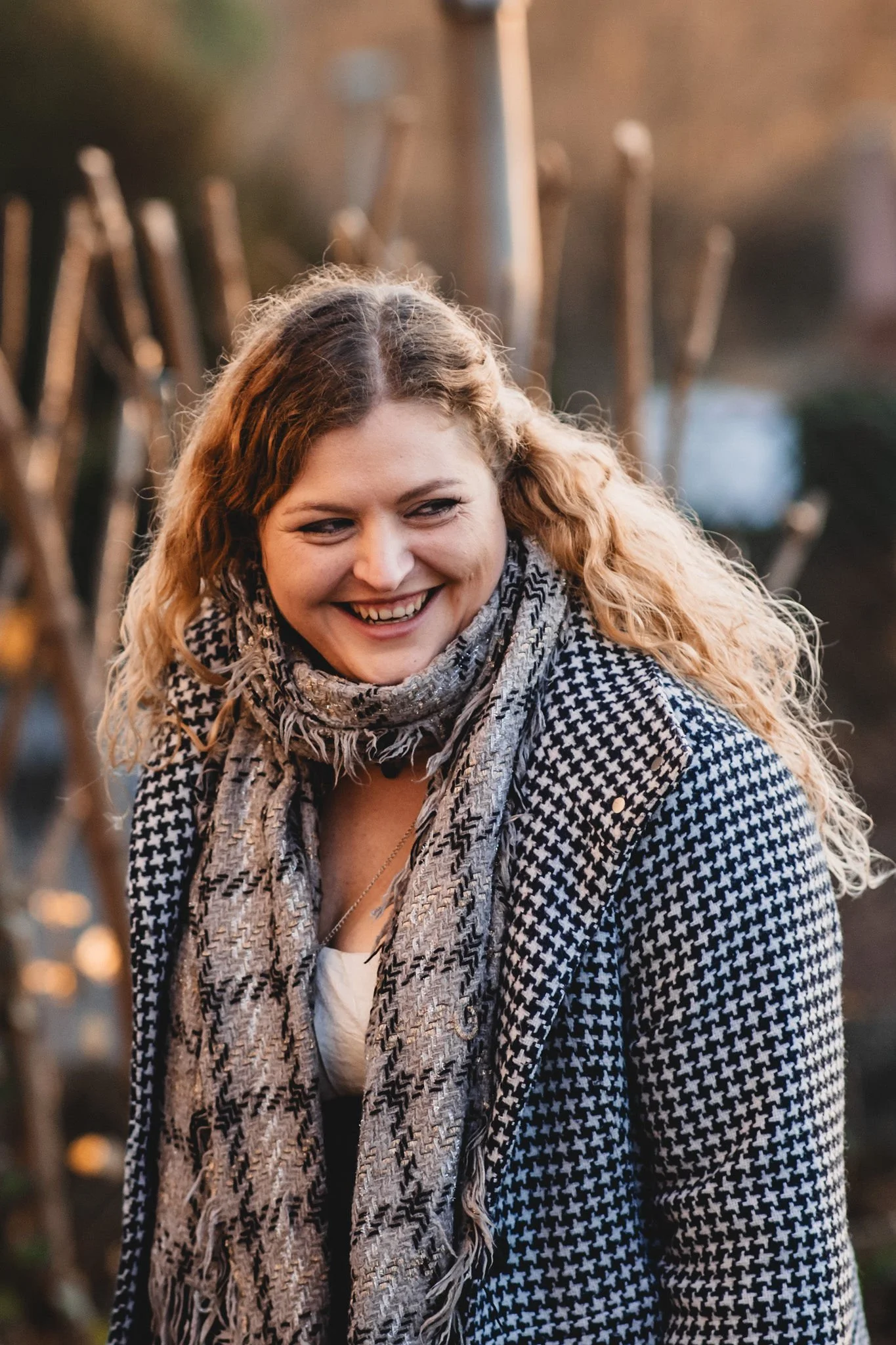 Eine lachende Frau mit lockigem, blondem Haar trägt einen schwarzen-weißen Hahnentritt-Blazer und einen grauen Schal, im Hintergrund ein stacheliger Zaun, bei Sonnenuntergang.