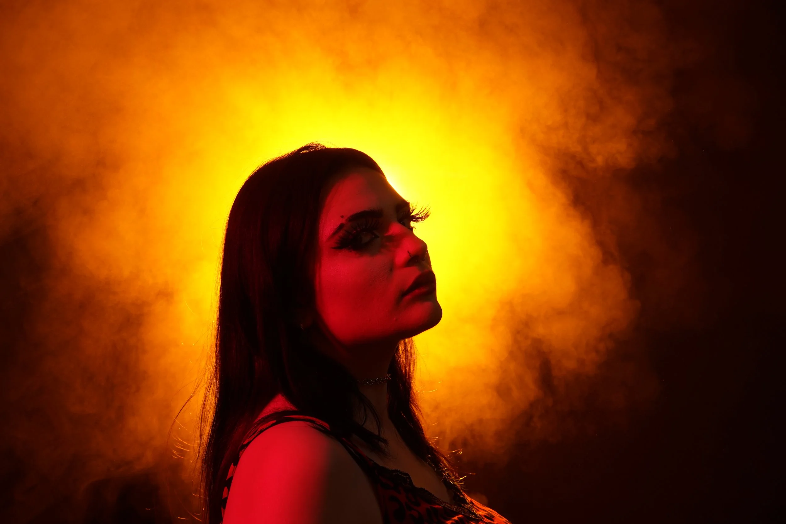 A woman with dark hair, dramatic makeup, and long eyelashes, facing left against a bright yellow and orange background.