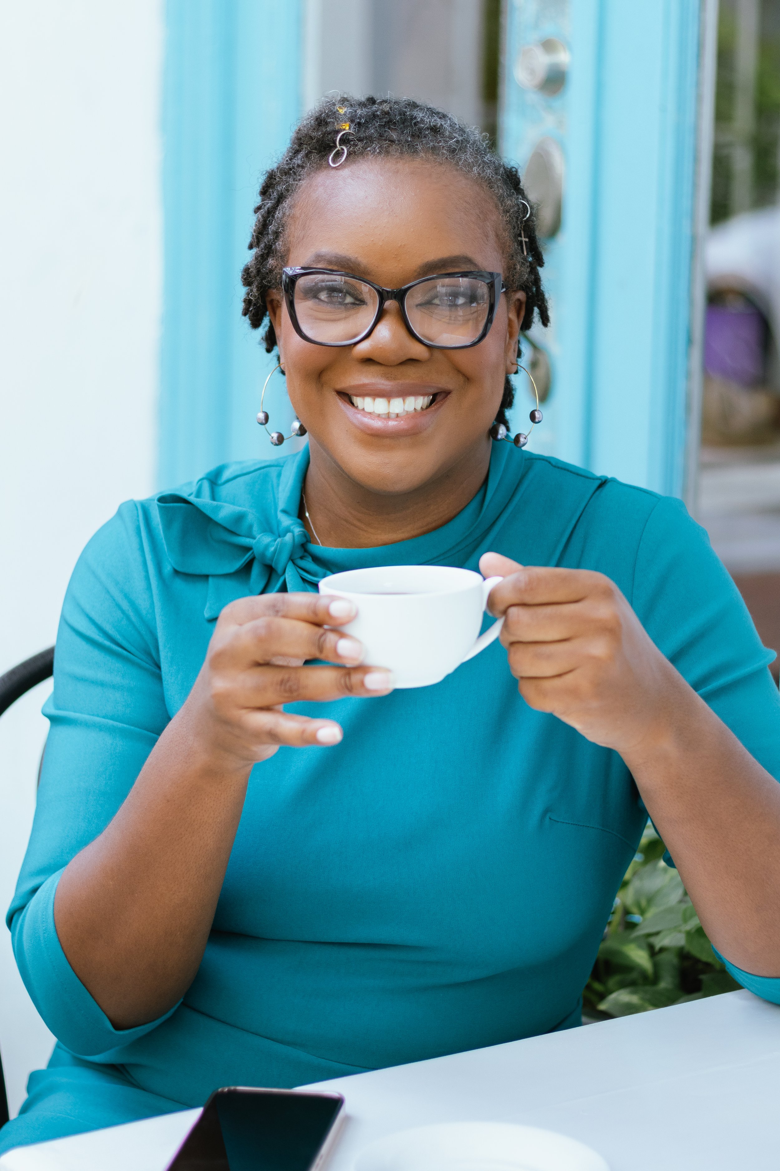 A woman with glasses and earrings smiling while holding a white coffee cup sitting outside at a table.