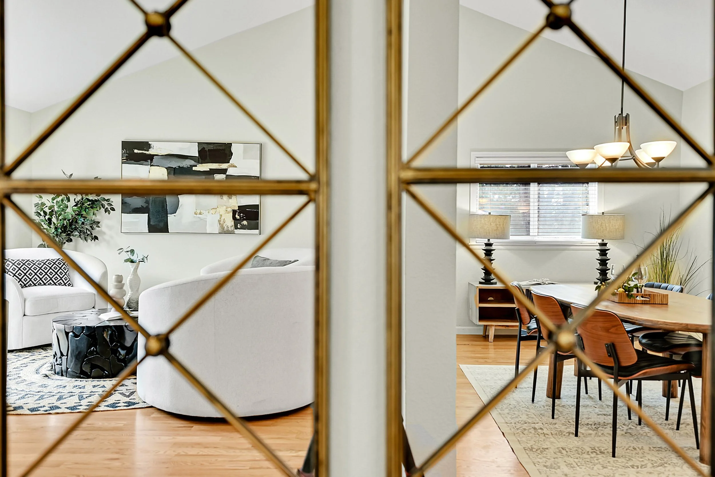View through decorative mirror showing a modern living room on the left with white sofas and abstract art on the wall, and a dining area on the right with a large wooden table, chairs, and lamps, with hardwood floors and a window with blinds.
