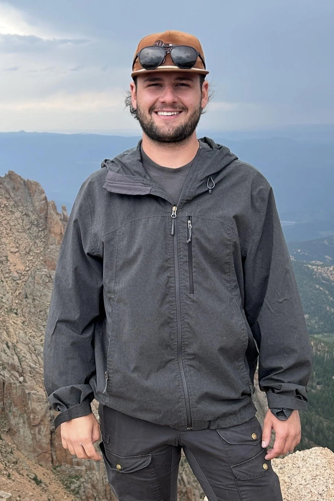 A man smiling while standing on a mountain with a scenic view of cliffs and mountains in the background, wearing a brown cap with sunglasses on top, a dark gray jacket, and black pants.
