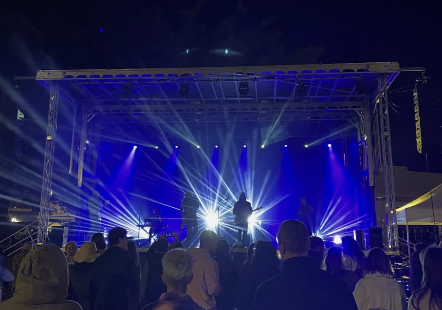 Live music performance on an outdoor stage at night, with bright blue and white stage lights, an audience watching, and silhouettes of band members playing instruments.