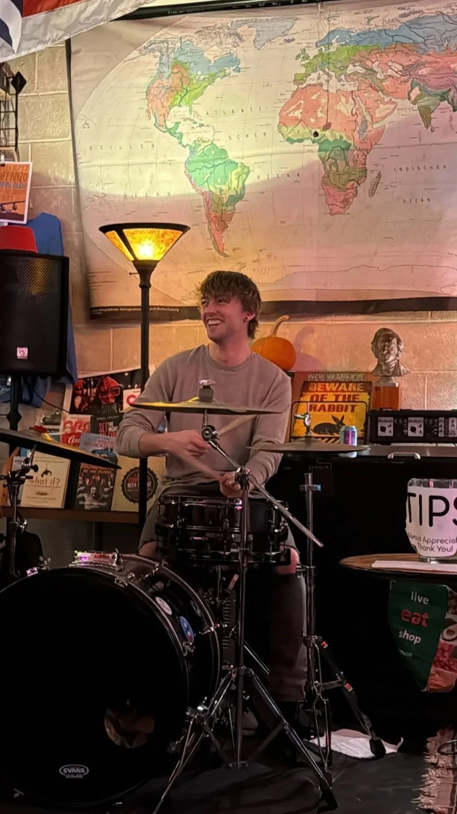 A young man is playing drums in a room decorated with various posters and objects, including a large world map on the wall, a pumpkin, a bust sculpture, and signs that say 'Beware of the Rabbit' and 'Tips Always Appreciated Thank You'.