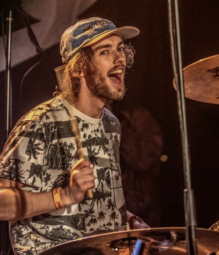 A young man playing drums at a live band performance, wearing a patterned shirt, a cap, and an orange wristband, with a cheerful expression.
