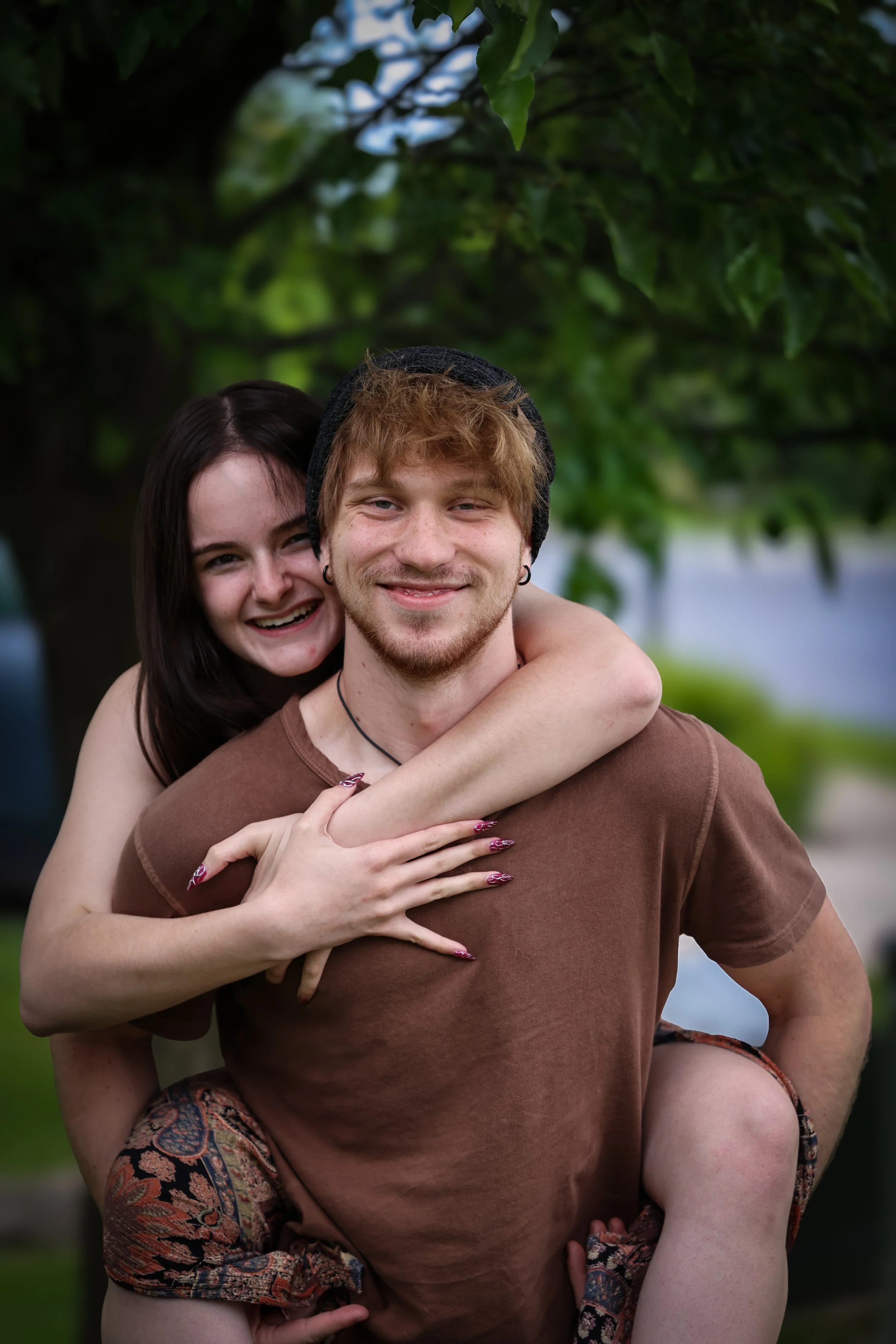 A smiling young woman with dark hair embracing a young man with light brown hair, a beard, and earrings, outdoors near a tree with green leaves.