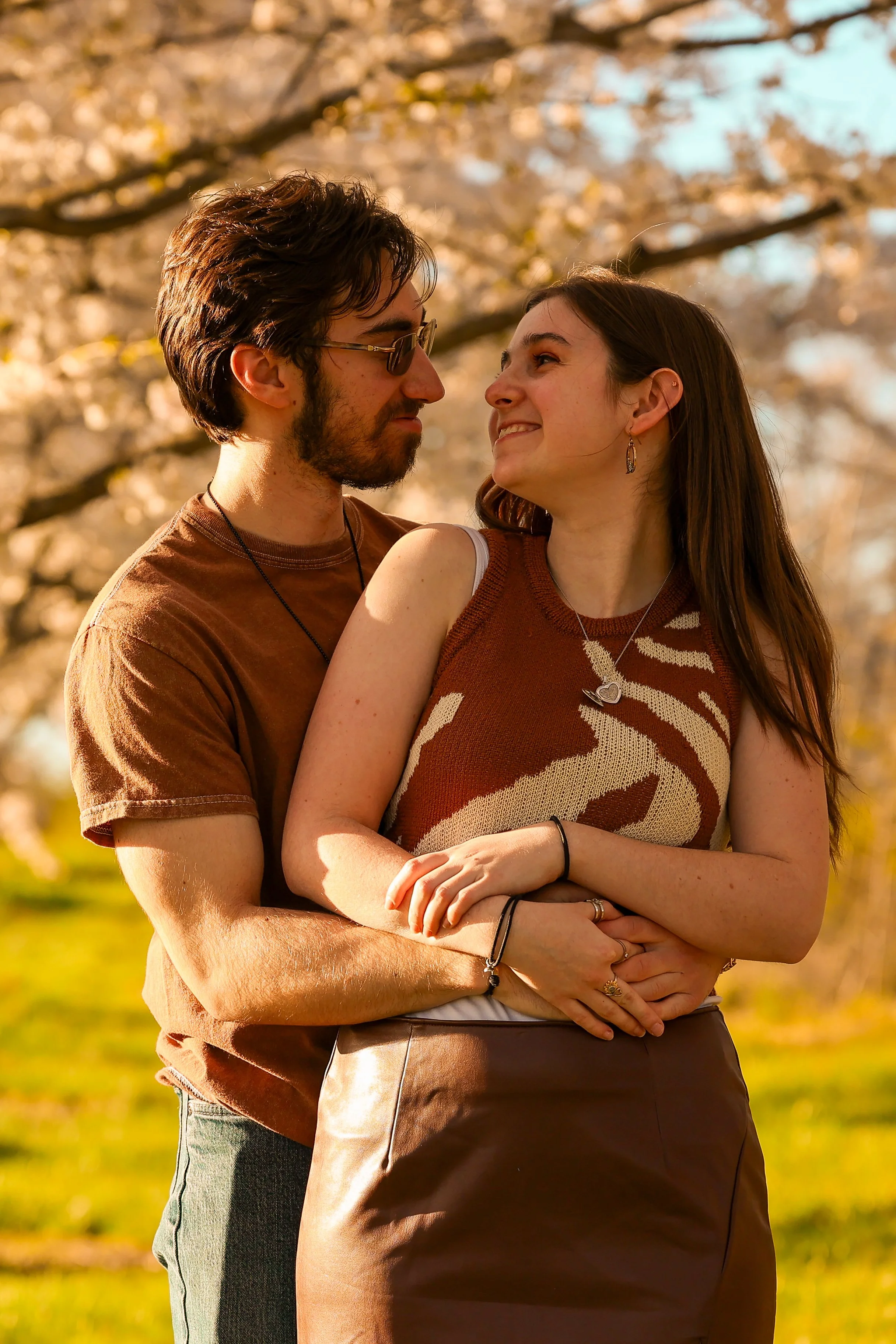 A couple is smiling and embracing outdoors in a park with autumn-colored trees in the background.