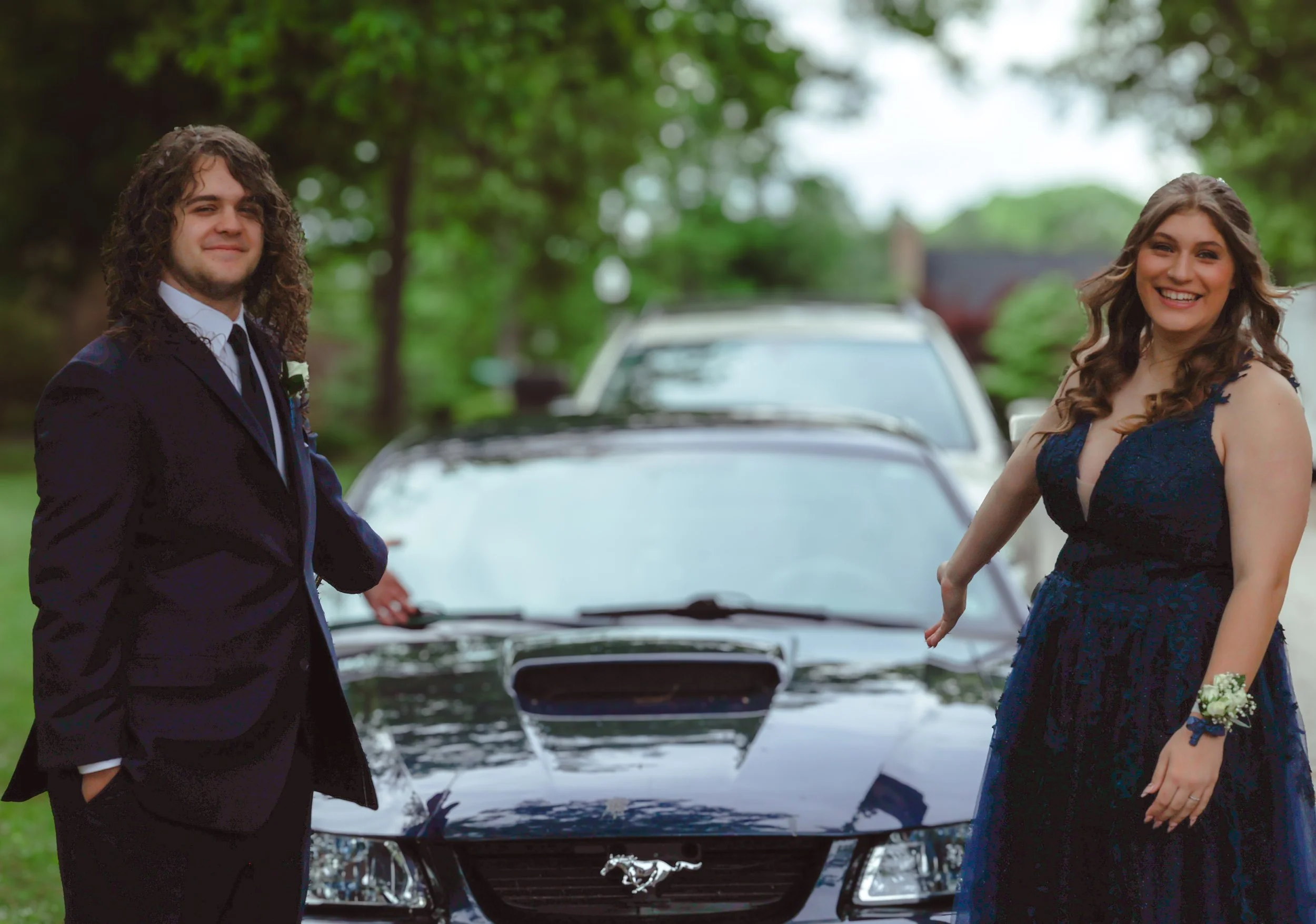 A bride and groom standing outdoors near a black car with a chrome grille, surrounded by greenery in a park-like setting. The groom has long curly hair, wearing a dark suit with a white shirt and tie. The bride has long wavy hair, wearing a dark blue