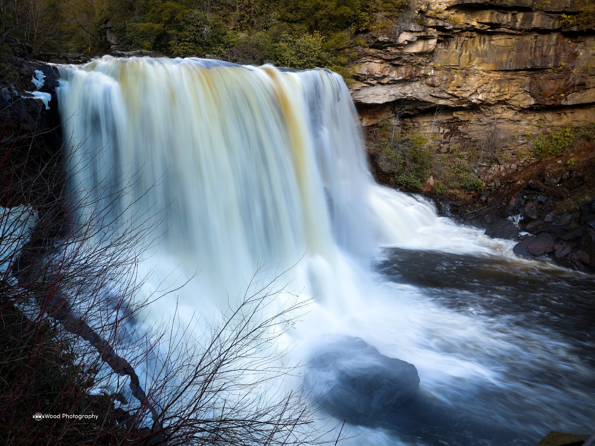 A large waterfall over rocks, surrounded by trees and cliffs, with white water rushing down into a pool below.