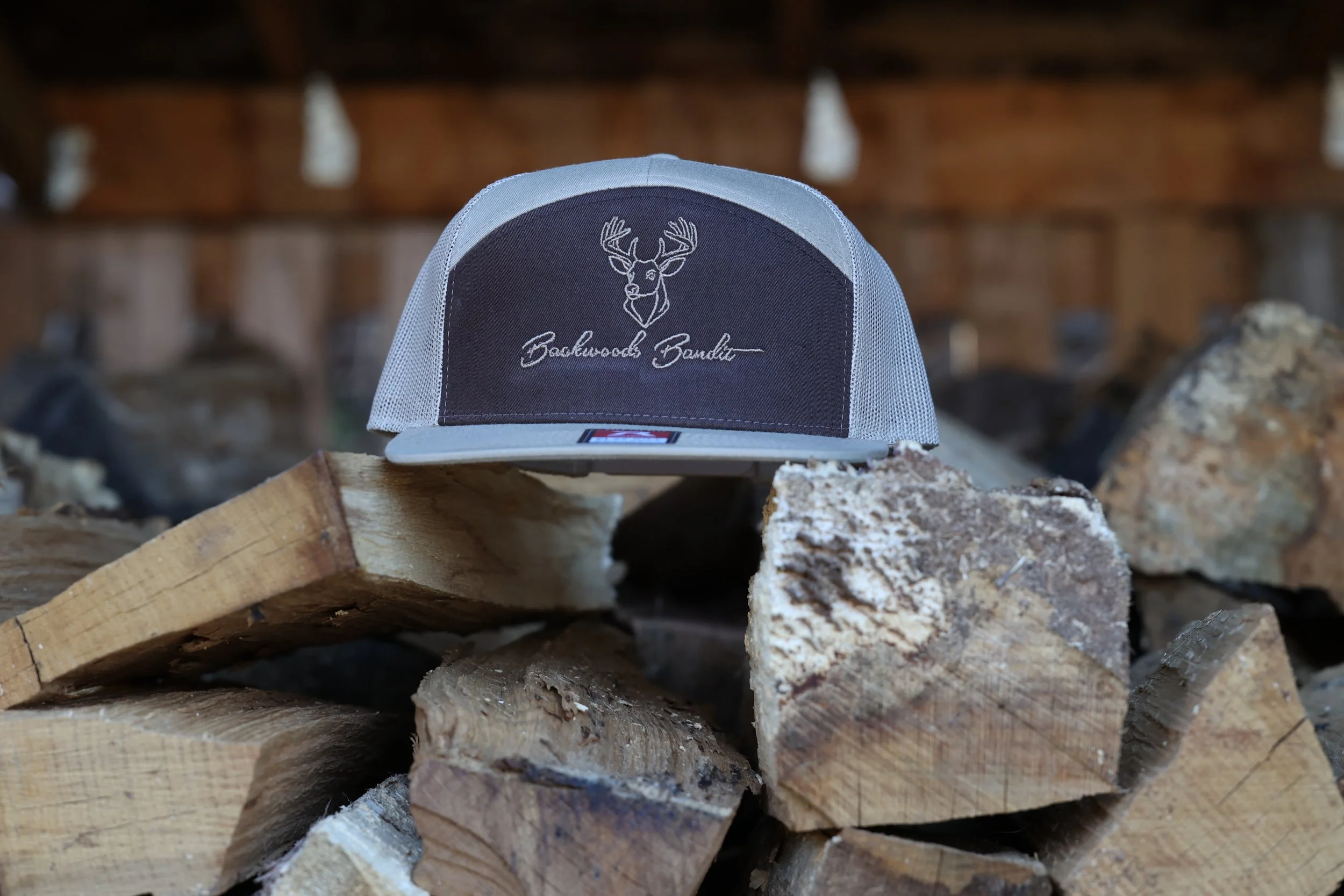 A gray and black trucker cap with a embroidered deer head and the text 'Backwoods Bandle' on the front, placed on stacked pieces of firewood.