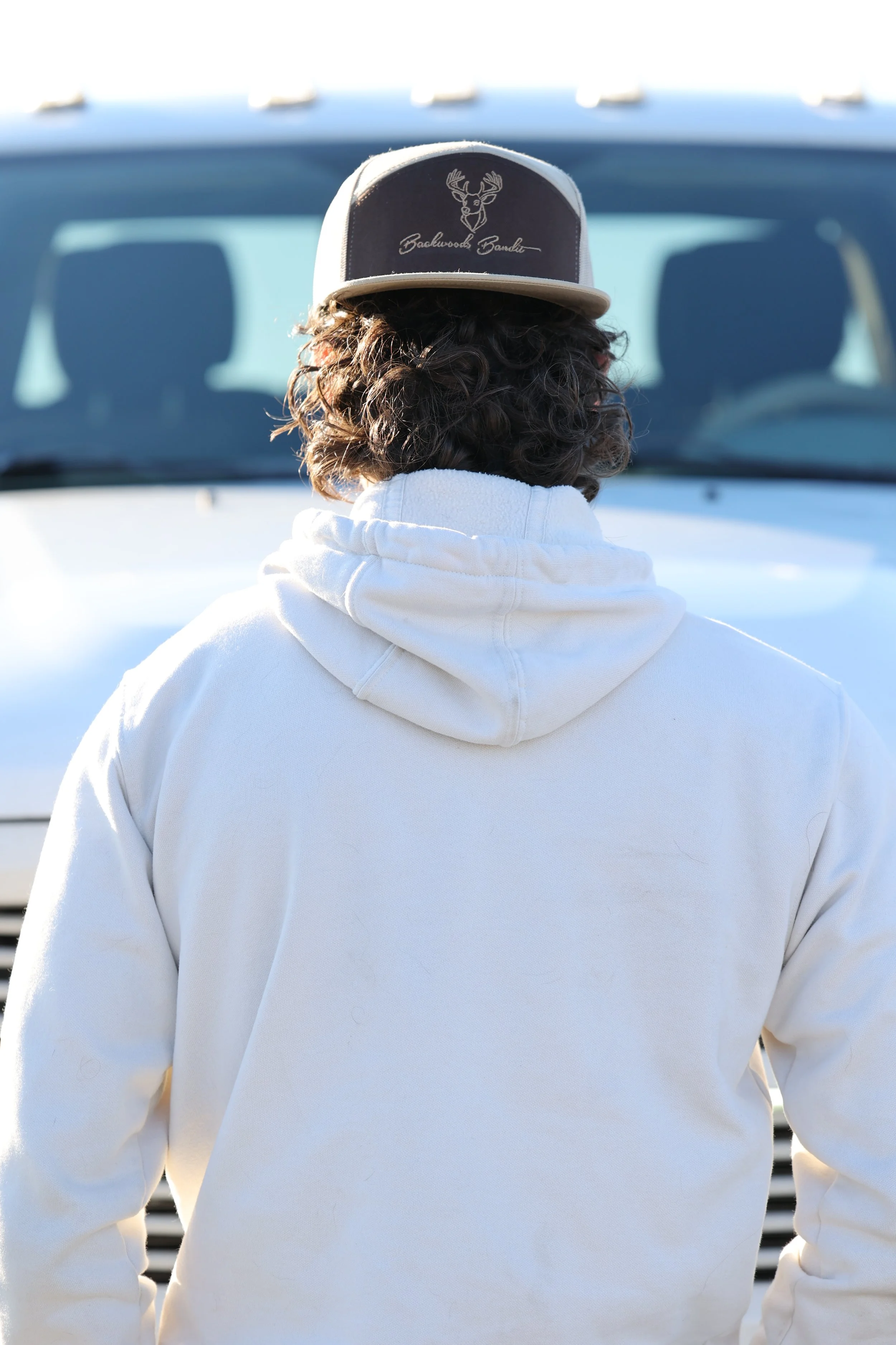 Person with curly hair wearing a white hoodie and a trucker hat with a deer logo and the words 'Backwoods Borduear,' facing away from the camera.