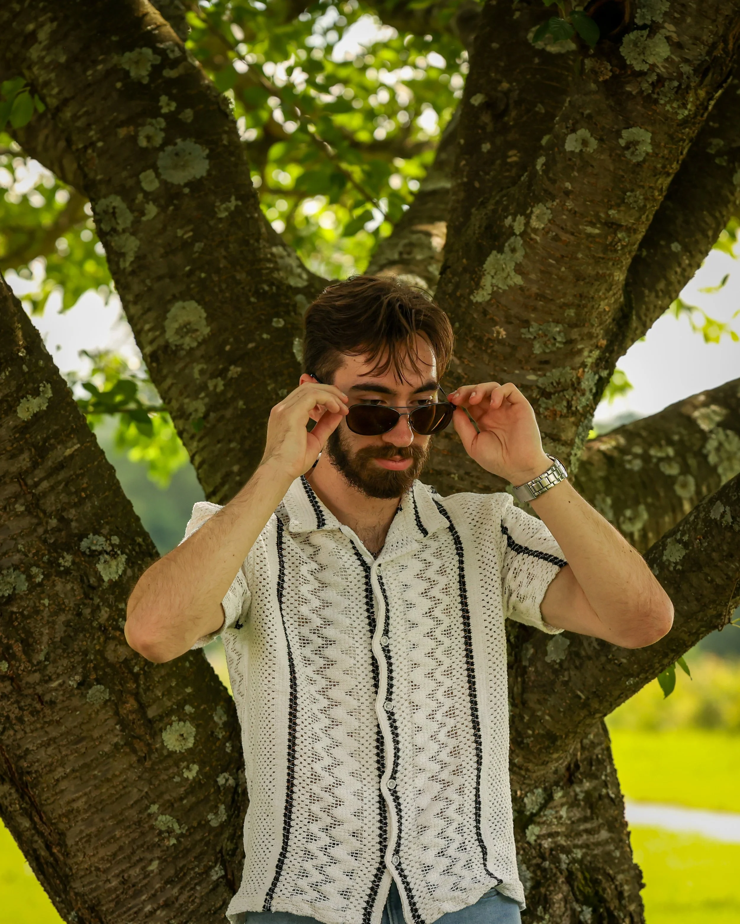 A man with a beard and wearing sunglasses, a watch, and a beige short-sleeved cardigan, stands in front of a large tree with green leaves, adjusting his sunglasses.
